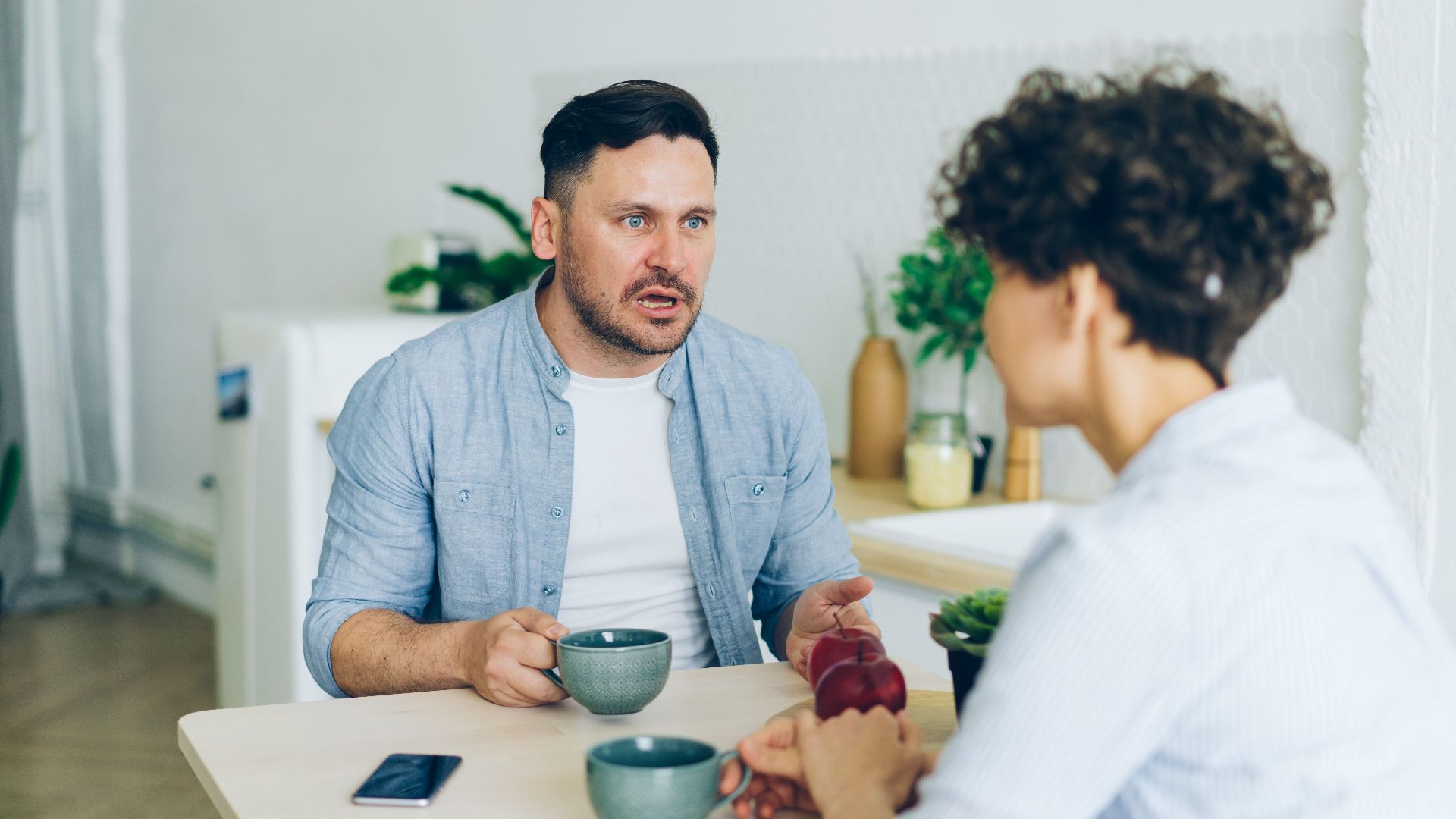 Couple having a serious conversation at the kitchen table with coffee cups.