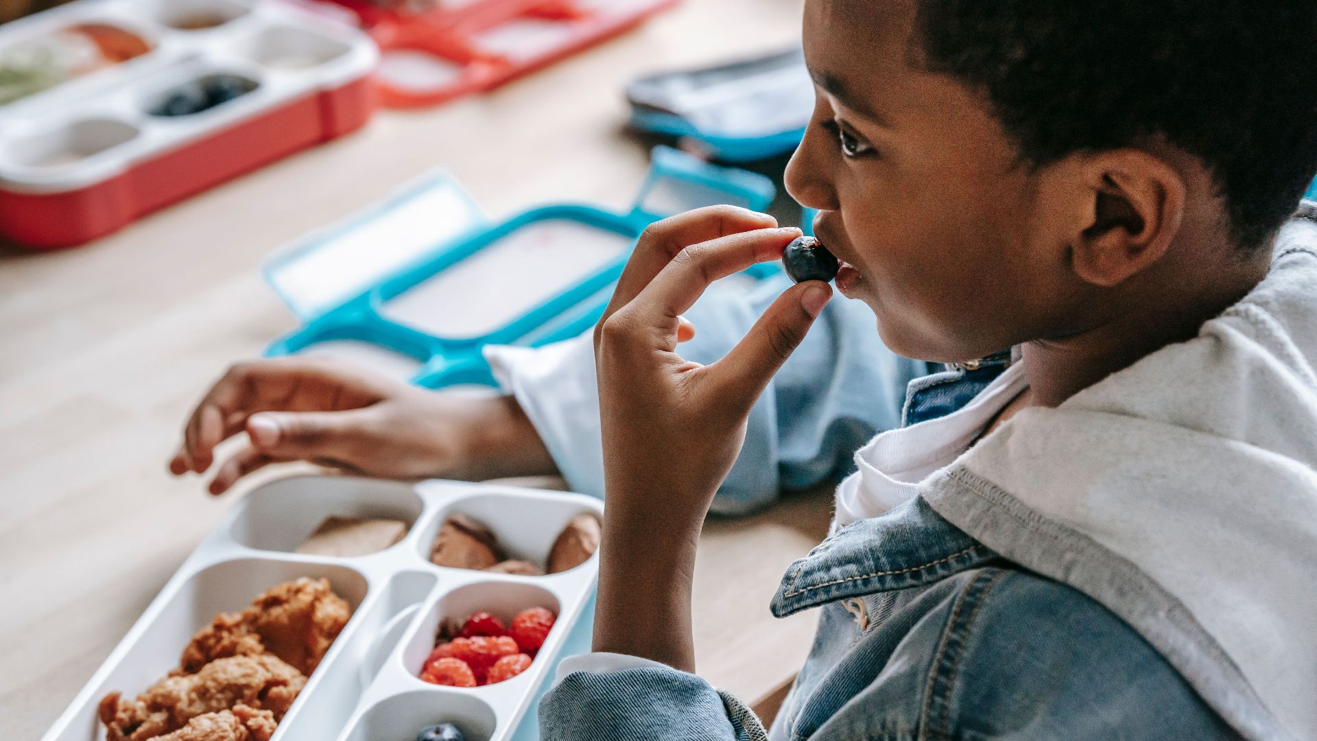 Crop African American child in casual clothes sitting at table with plastic lunch box while having snack