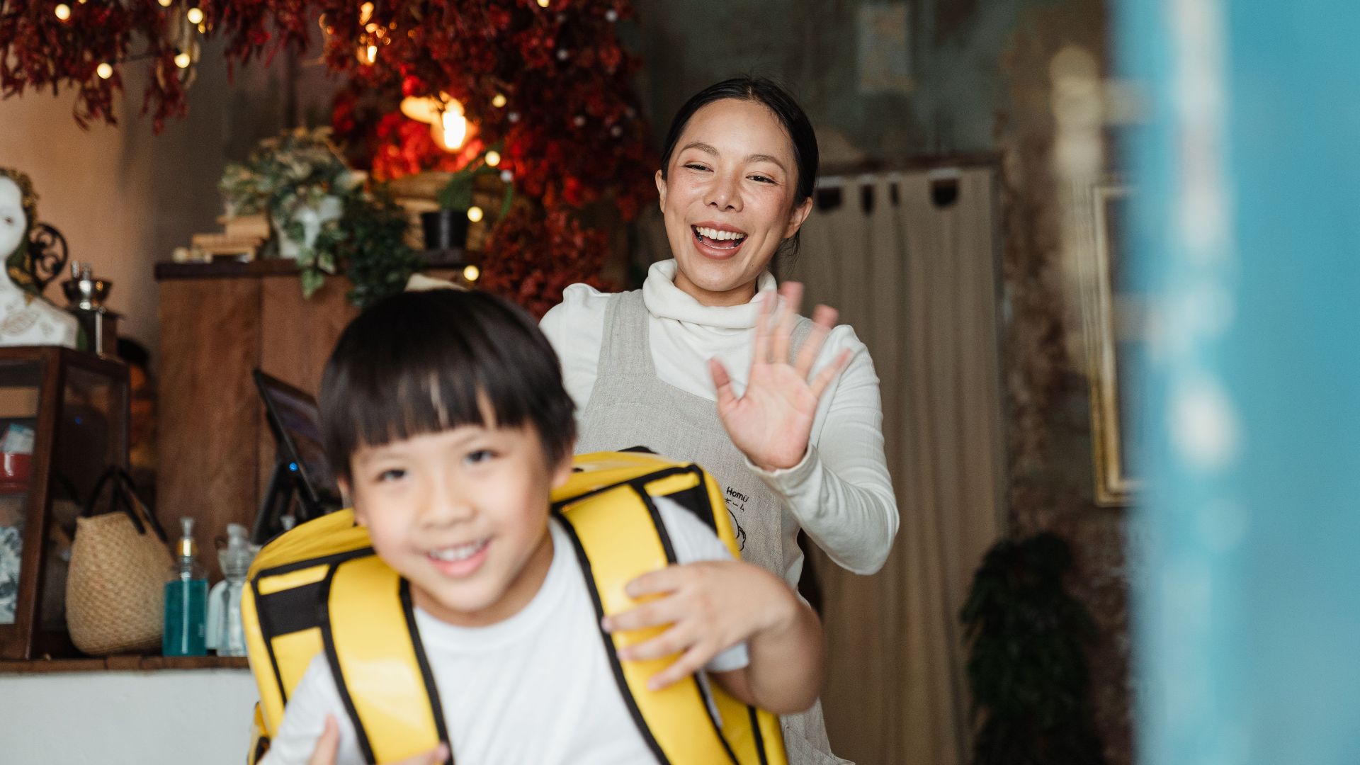Content ethnic woman in apron showing bye gesture to positive boy with yellow food rucksack looking away at home decorated with garland