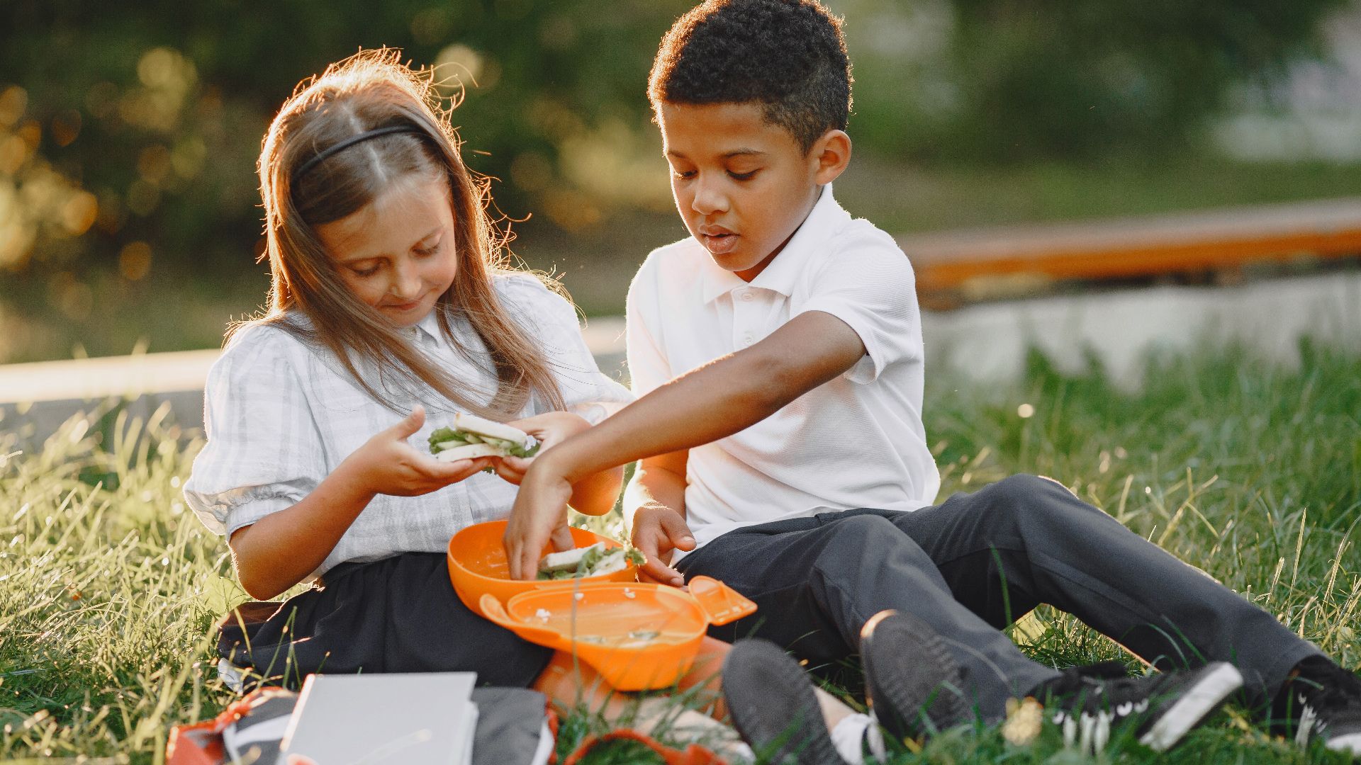 Two children in school uniforms enjoy lunch together outside, sharing sandwiches from a lunchbox.