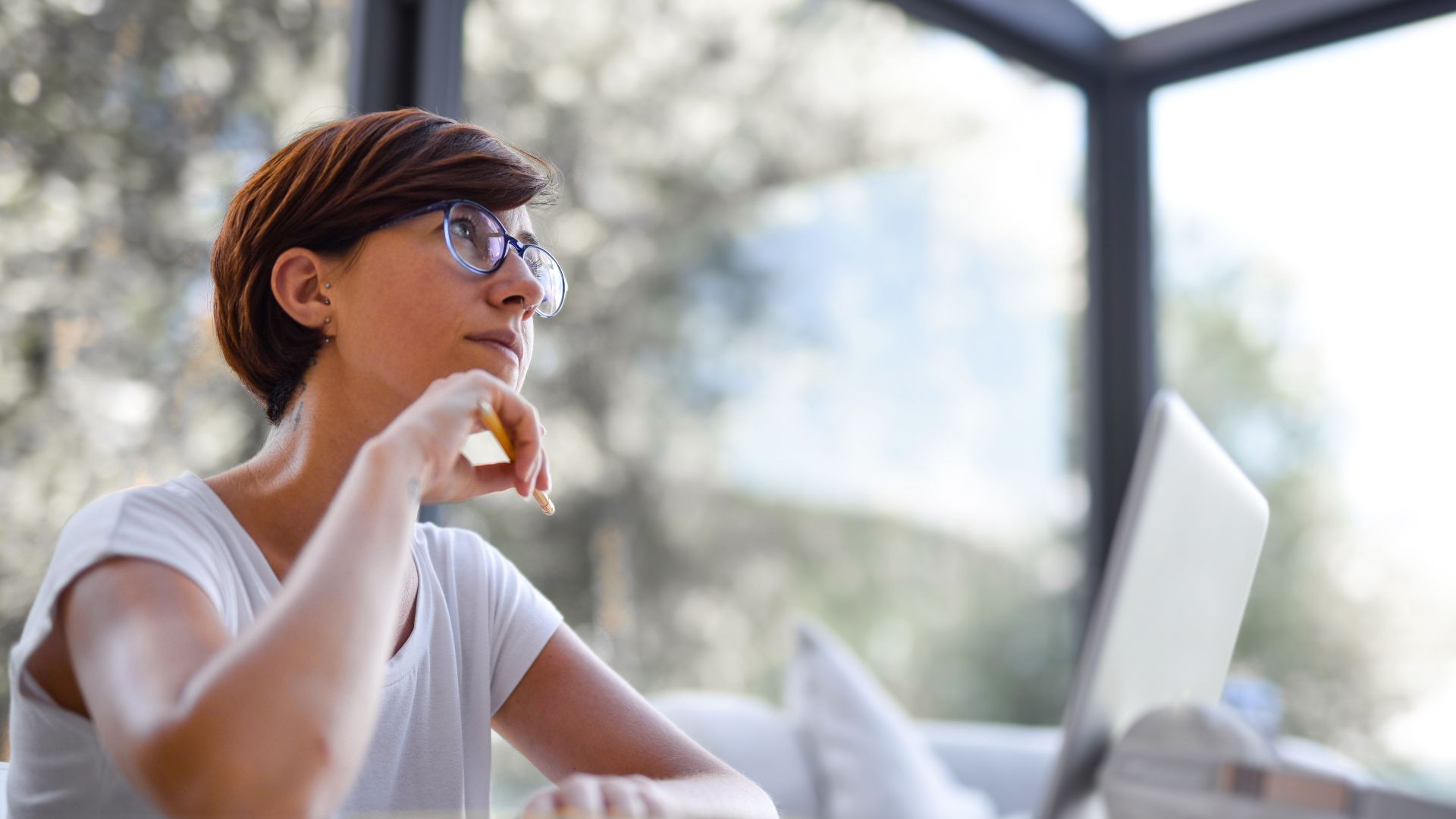 Thoughtful woman with glasses and short hair looking out window in a modern room.