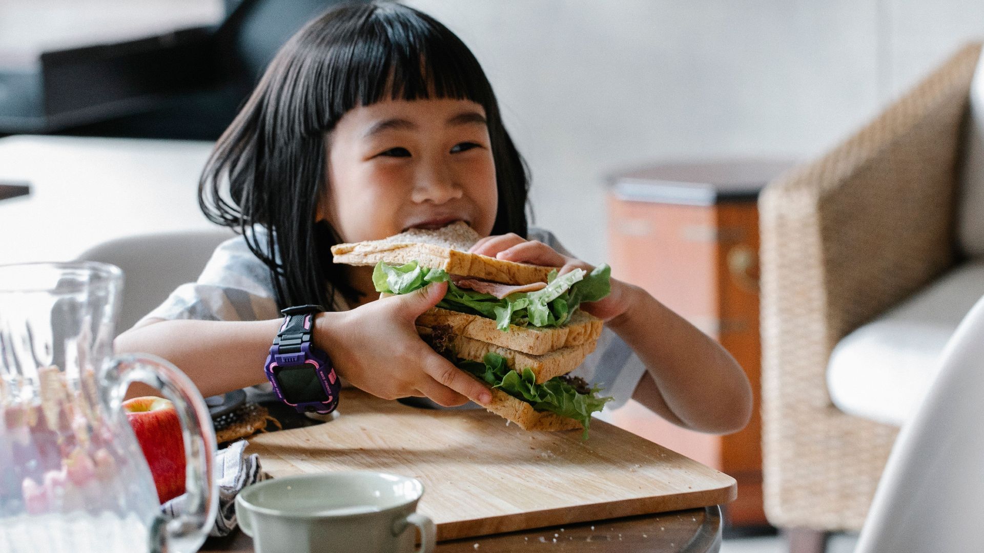 Asian child sitting at wooden table during breakfast and eating tasty sandwich with lettuce leaves