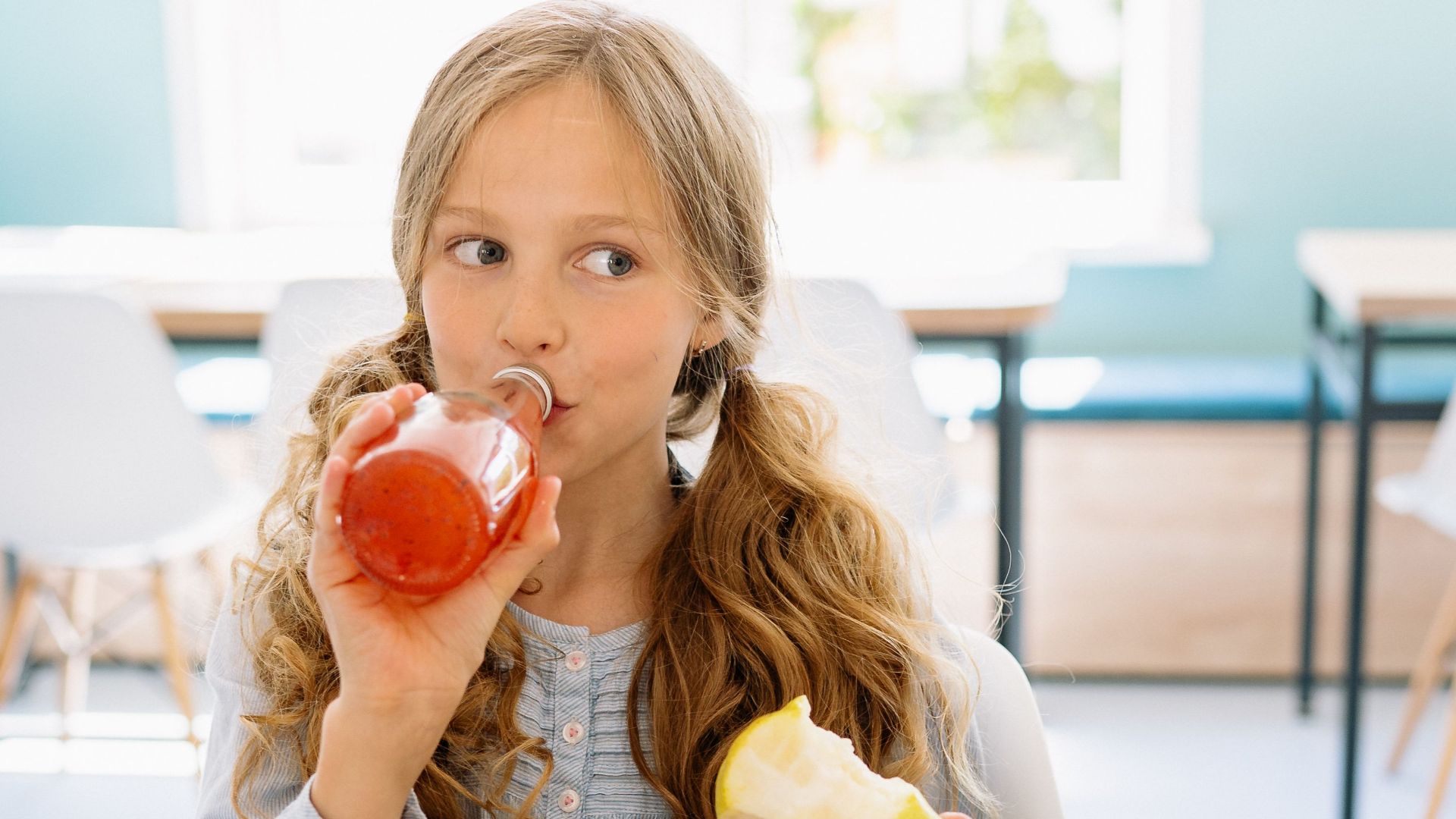 A young girl drinks a beverage and eats an apple during a break in a classroom setting.