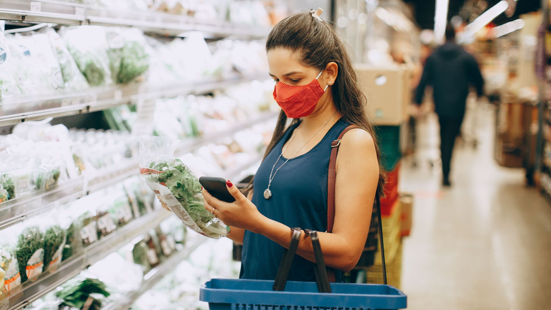 A woman wearing a mask shops for groceries in a supermarket, checking products with her phone.