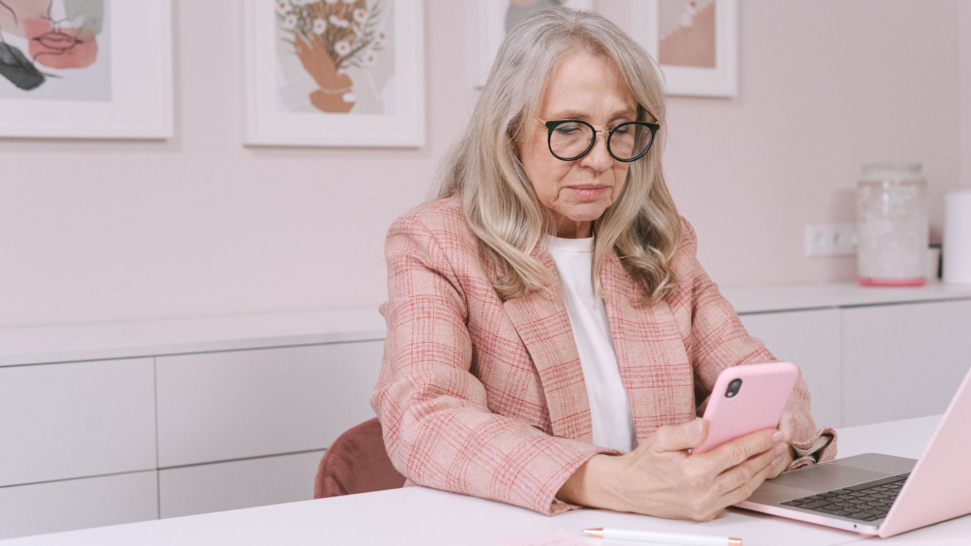 Elderly woman in pink blazer using smartphone and laptop, working remotely from a stylish workspace.