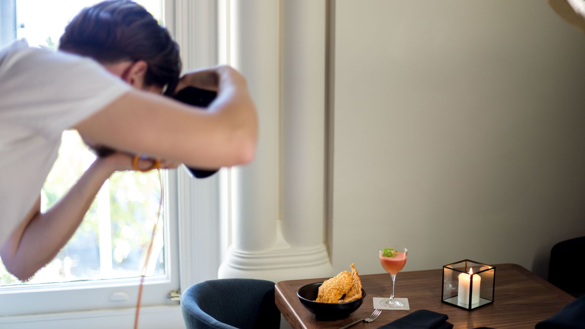 A photographer taking pictures of a stylish table with food and drinks by the window.