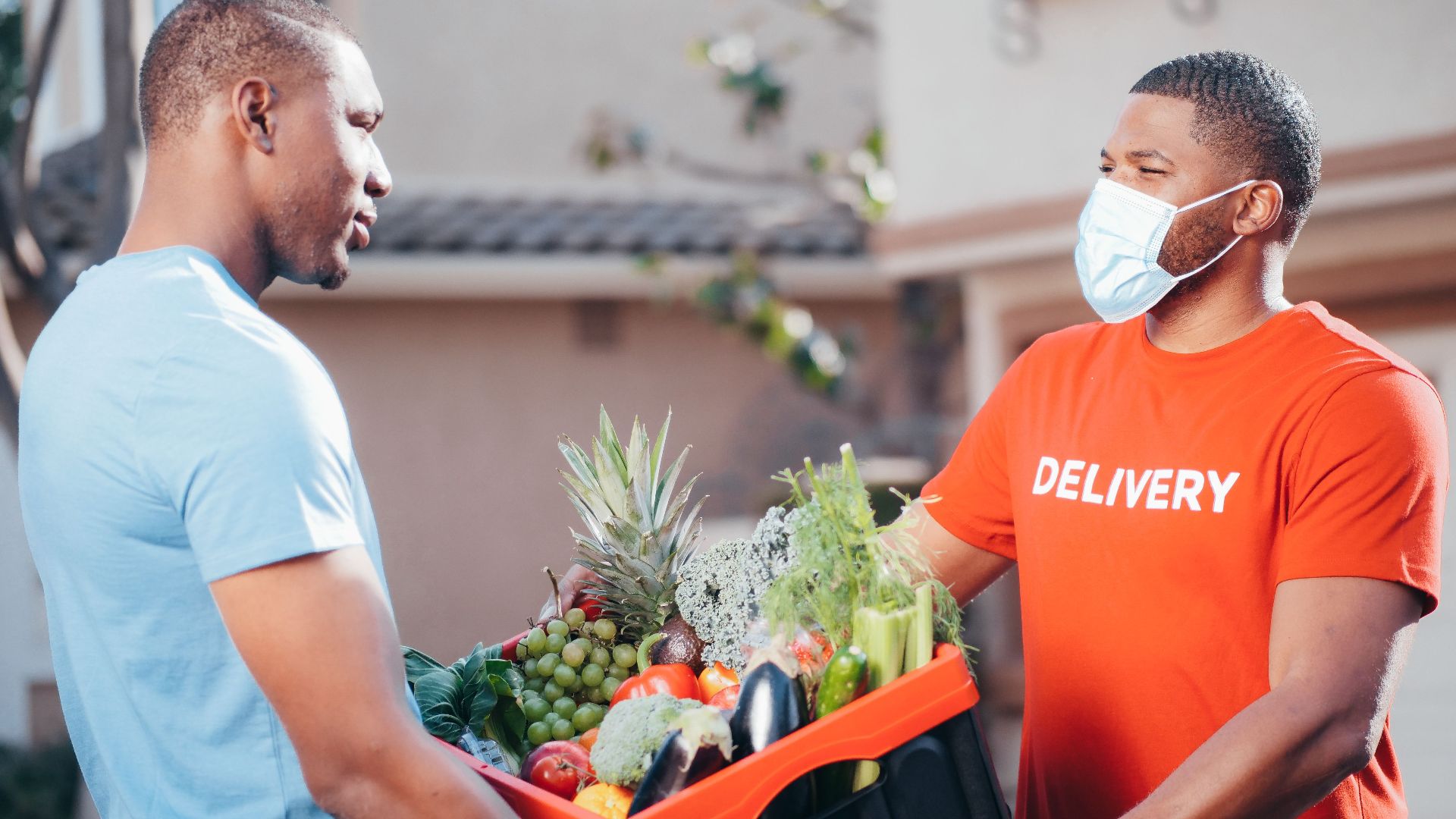 Two men exchanging a box of fresh produce for contactless delivery in a residential area.