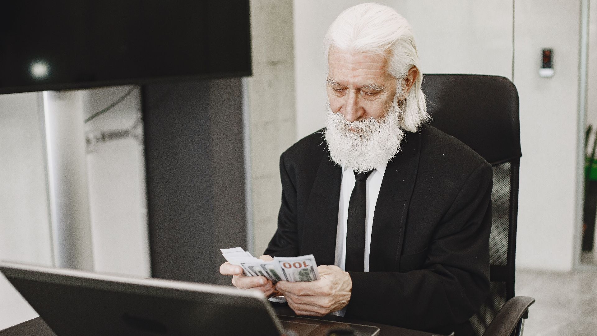 Senior businessman in suit counting money at office desk with a laptop. Elegance and financial focus in modern workspace.