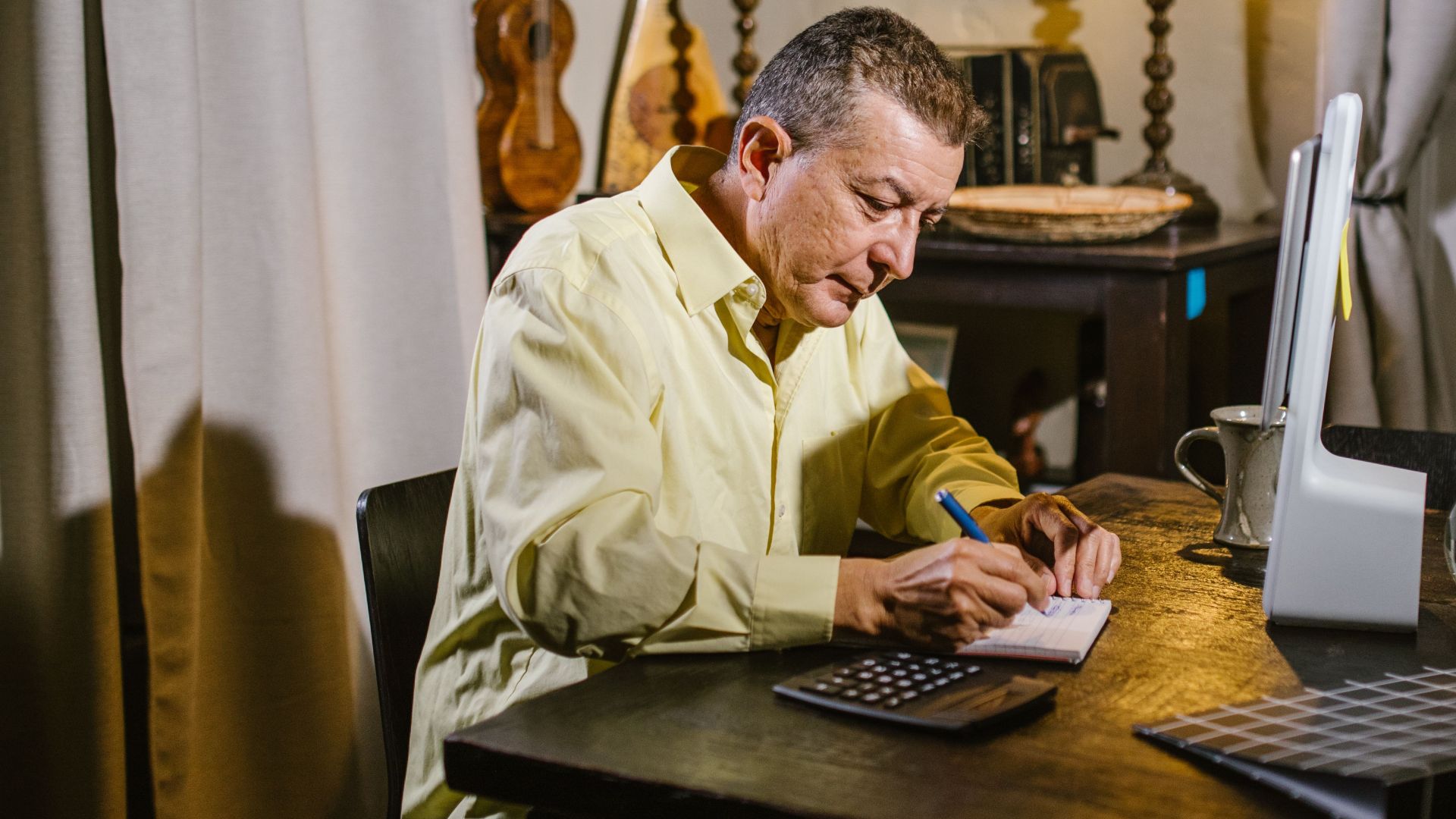 Senior man wearing yellow shirt writing notes at desk with lamp, calculator, and glass sphere nearby.