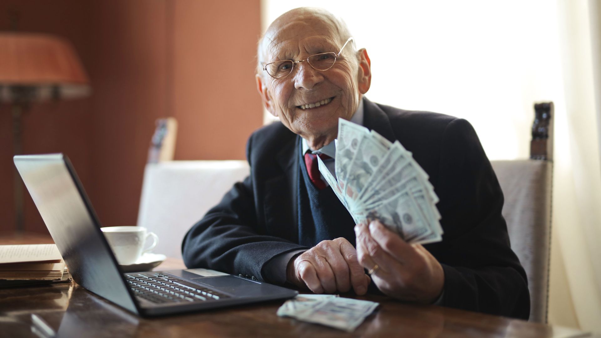 Positive senior businessman in formal suit and eyeglasses holding bunch of bills while working on laptop at table and looking at camera