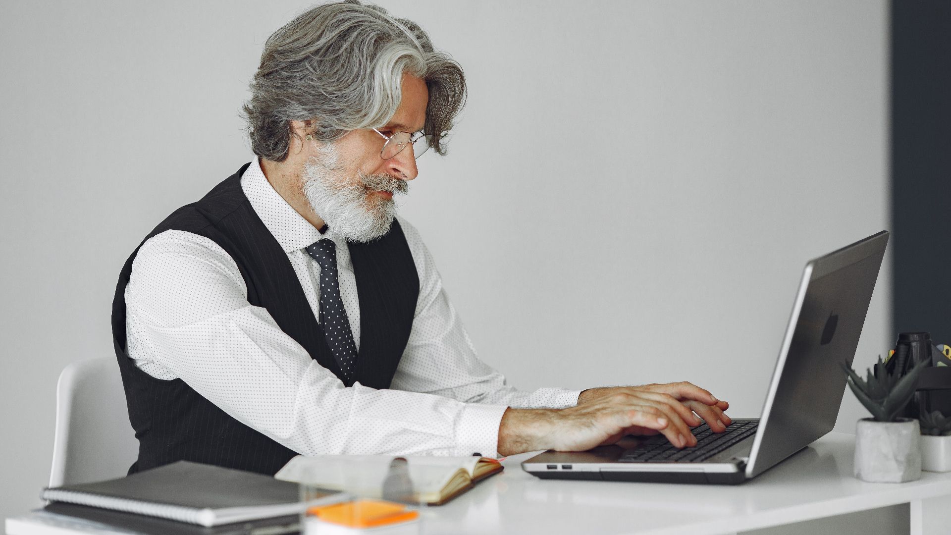 Elegant senior man in office attire typing on a laptop at a desk.