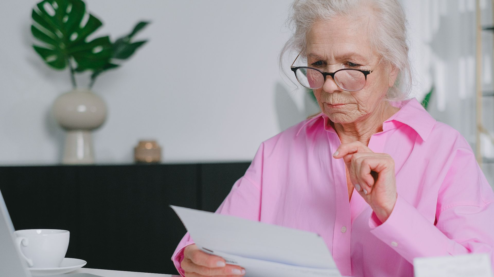 Elderly woman in pink shirt reading documents at home workspace.