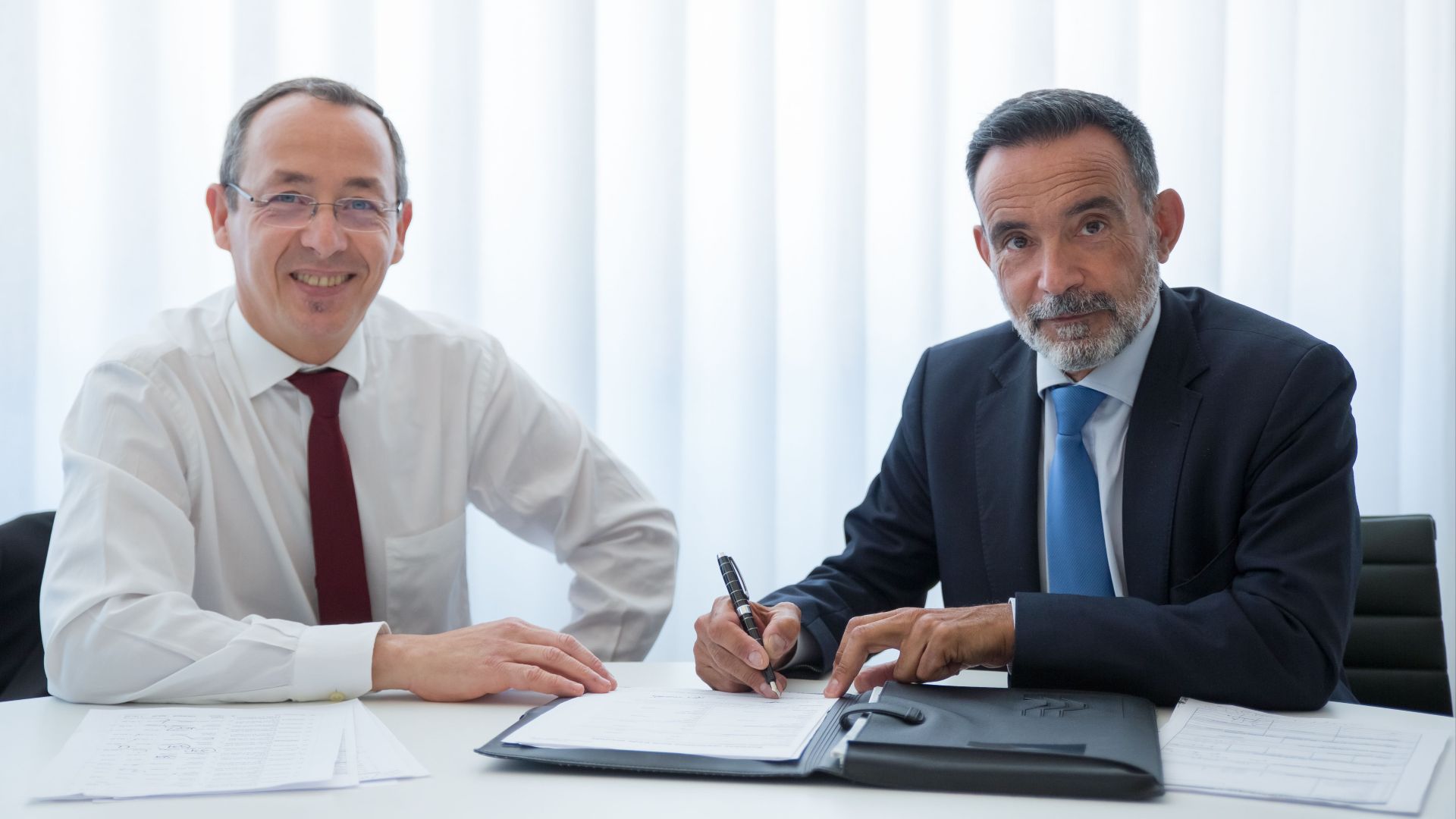 Two businessmen discussing and signing documents at a bright office table.