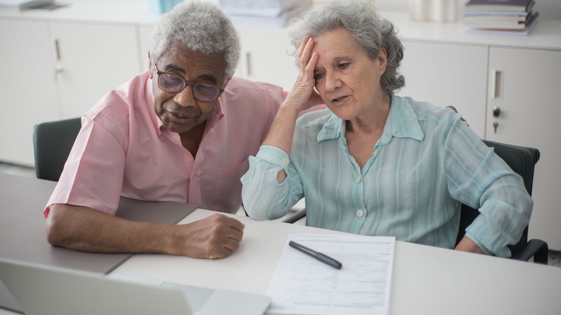 Elderly couple engaged with a laptop, reviewing paperwork at home office desk.