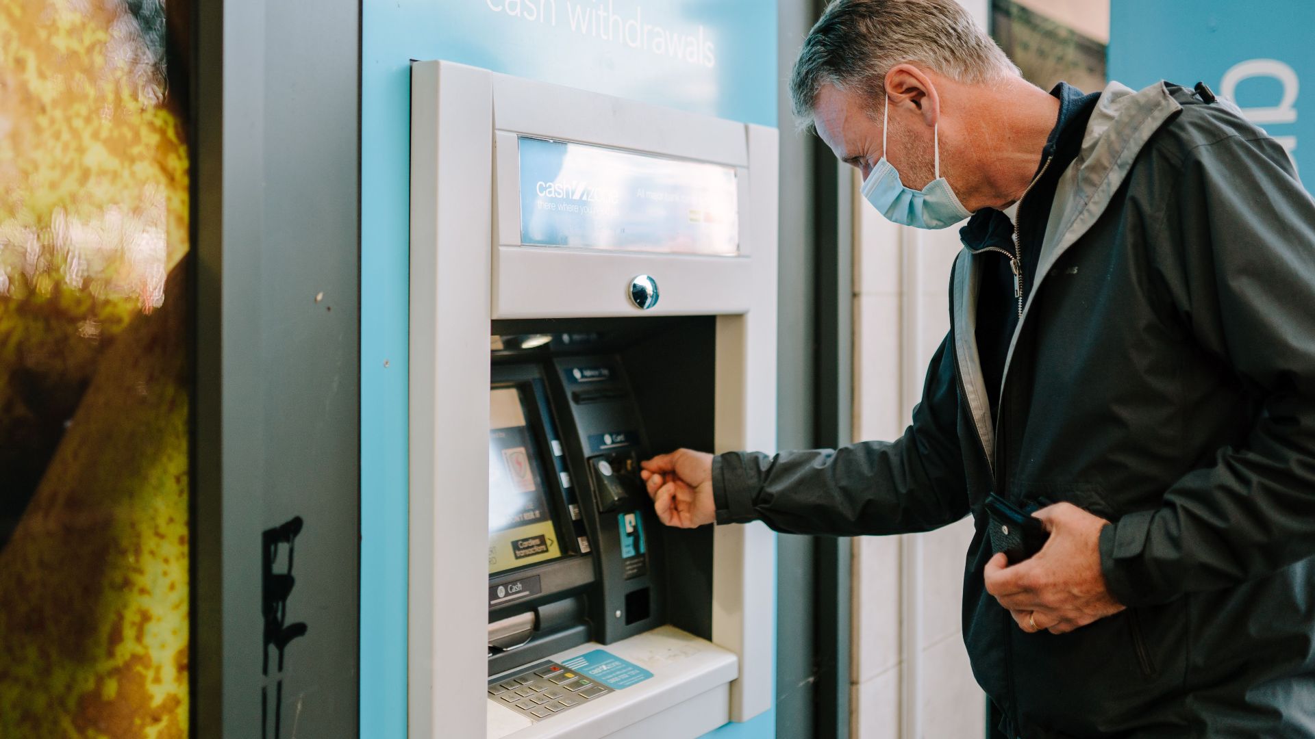 A man wearing a mask using an ATM machine outdoors for cash withdrawal.