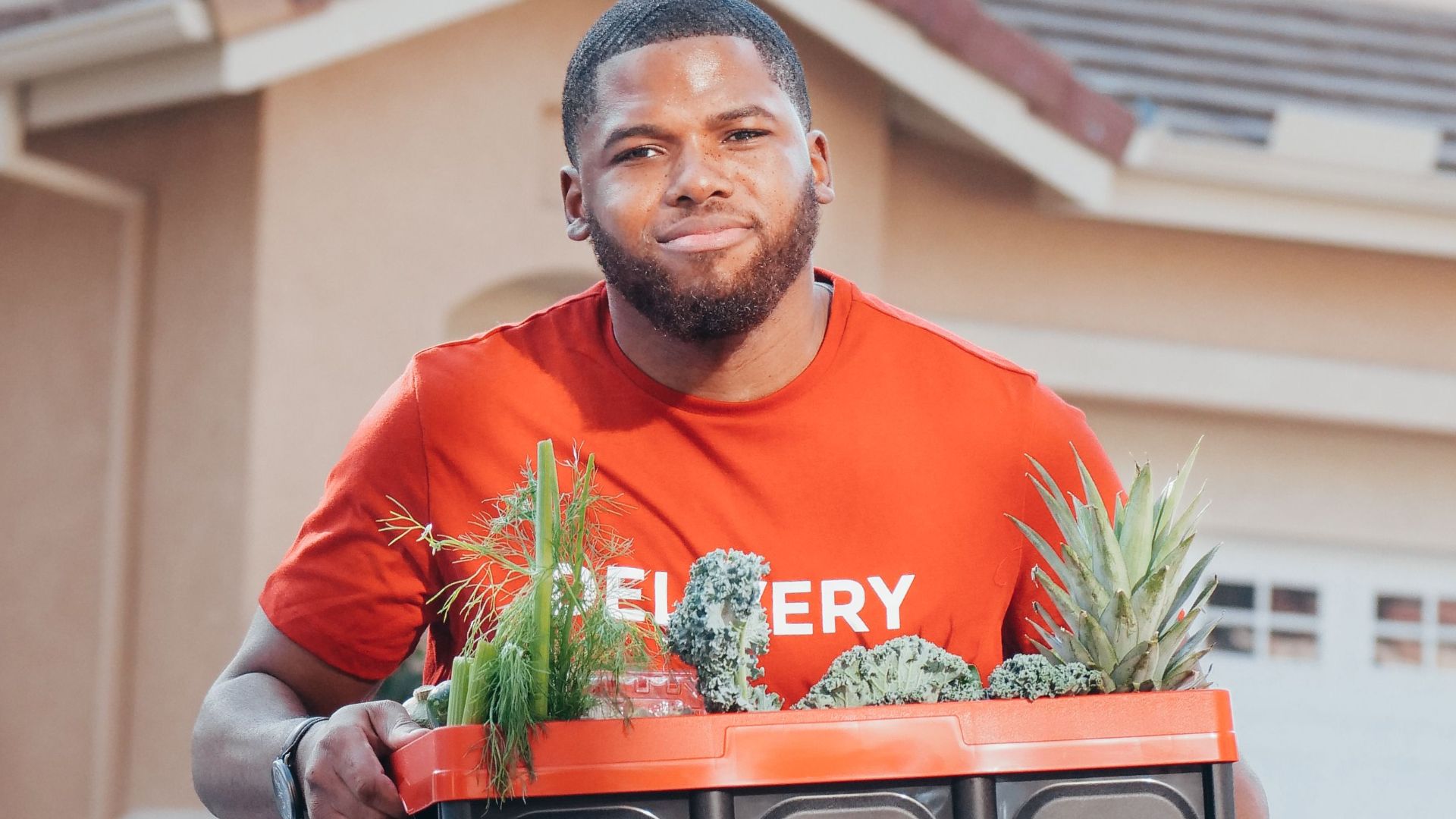 Delivery man carrying a box of groceries with pineapples outside a house.