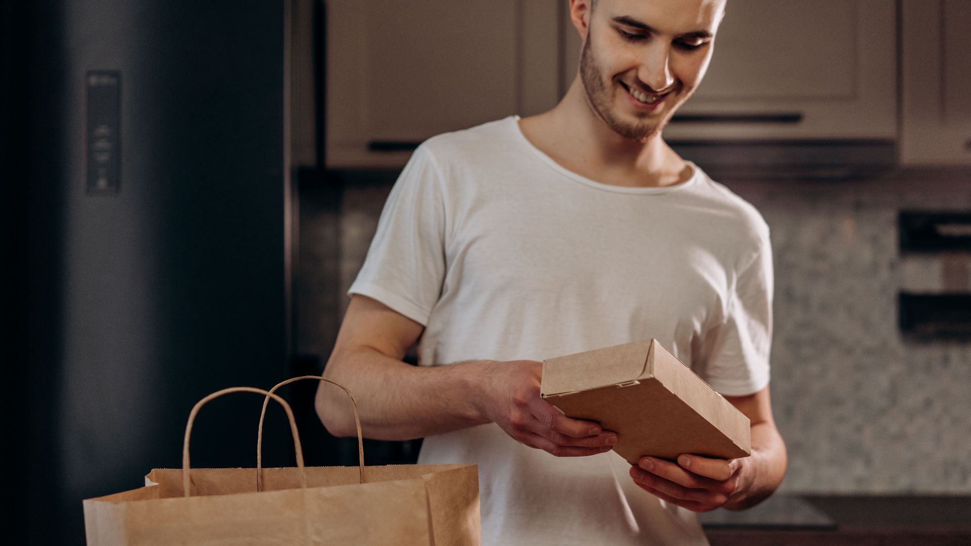 A man happily unpacks a food delivery in a modern kitchen setting.
