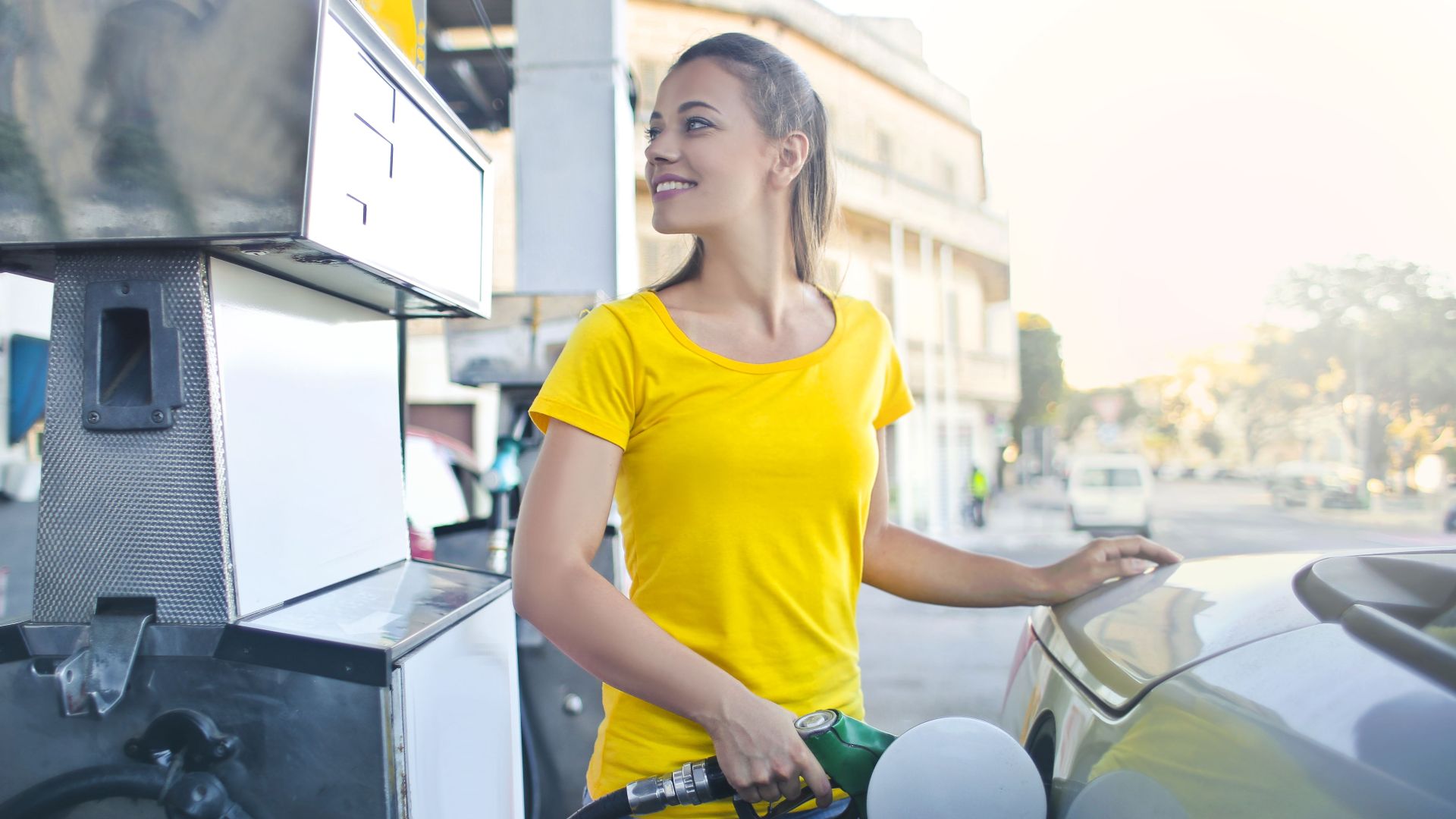 A smiling woman in a yellow shirt refuels her car at a gas station, enjoying a sunny day.