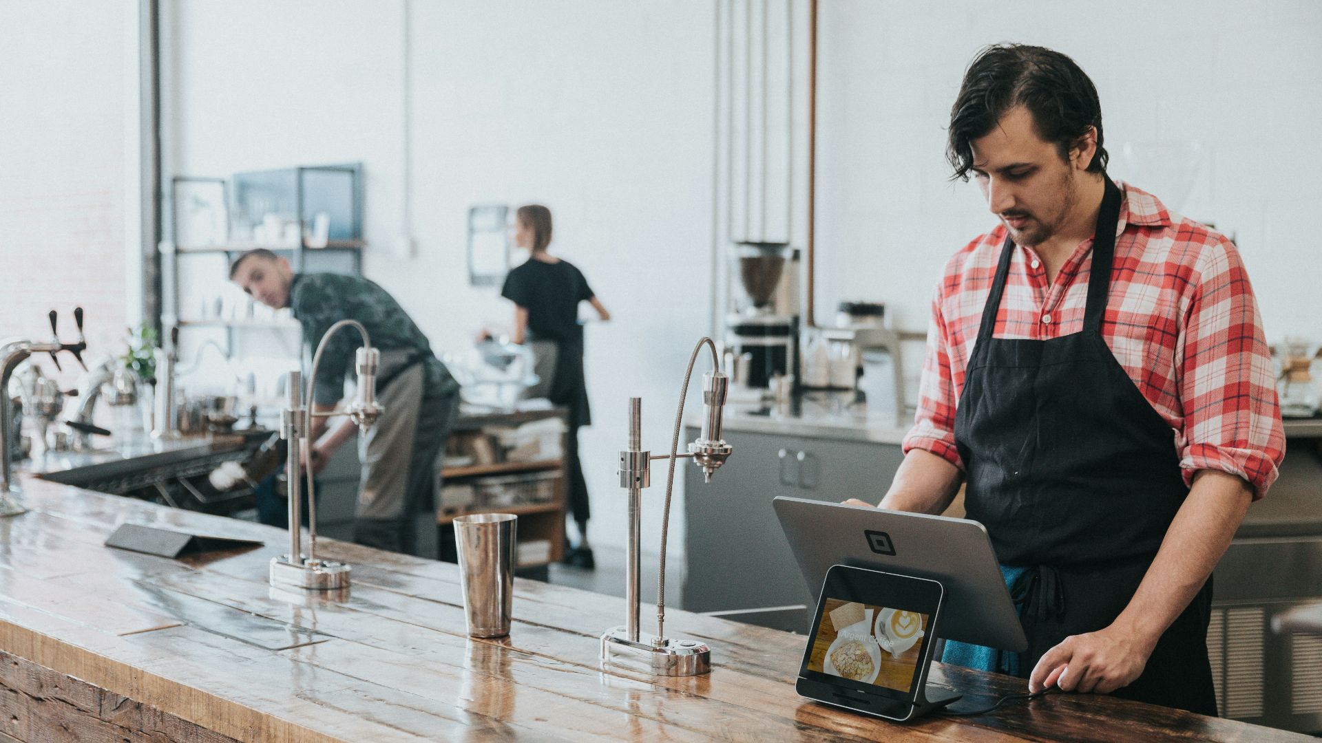 A barista in a plaid shirt and apron using a tablet at a wooden counter