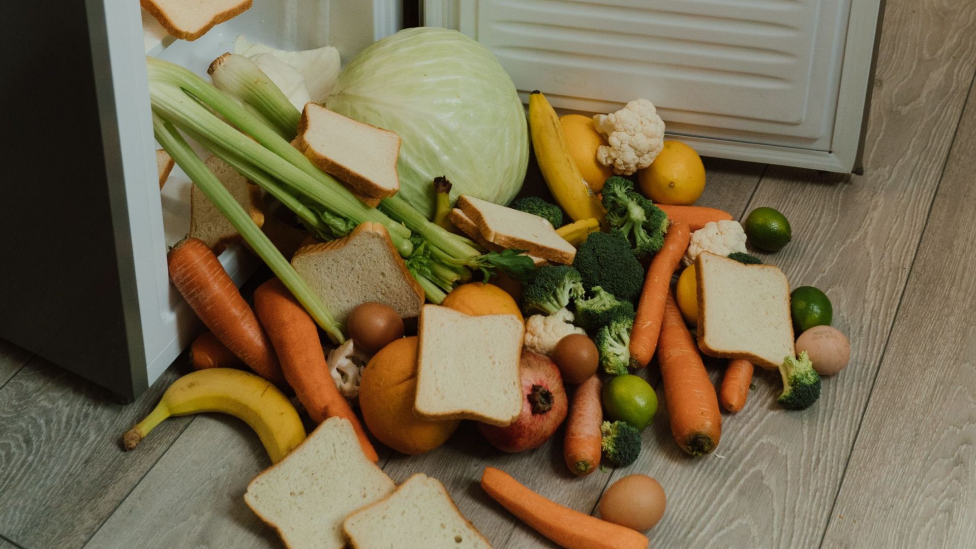 A messy open refrigerator with fresh vegetables and bread scattered on the floor.