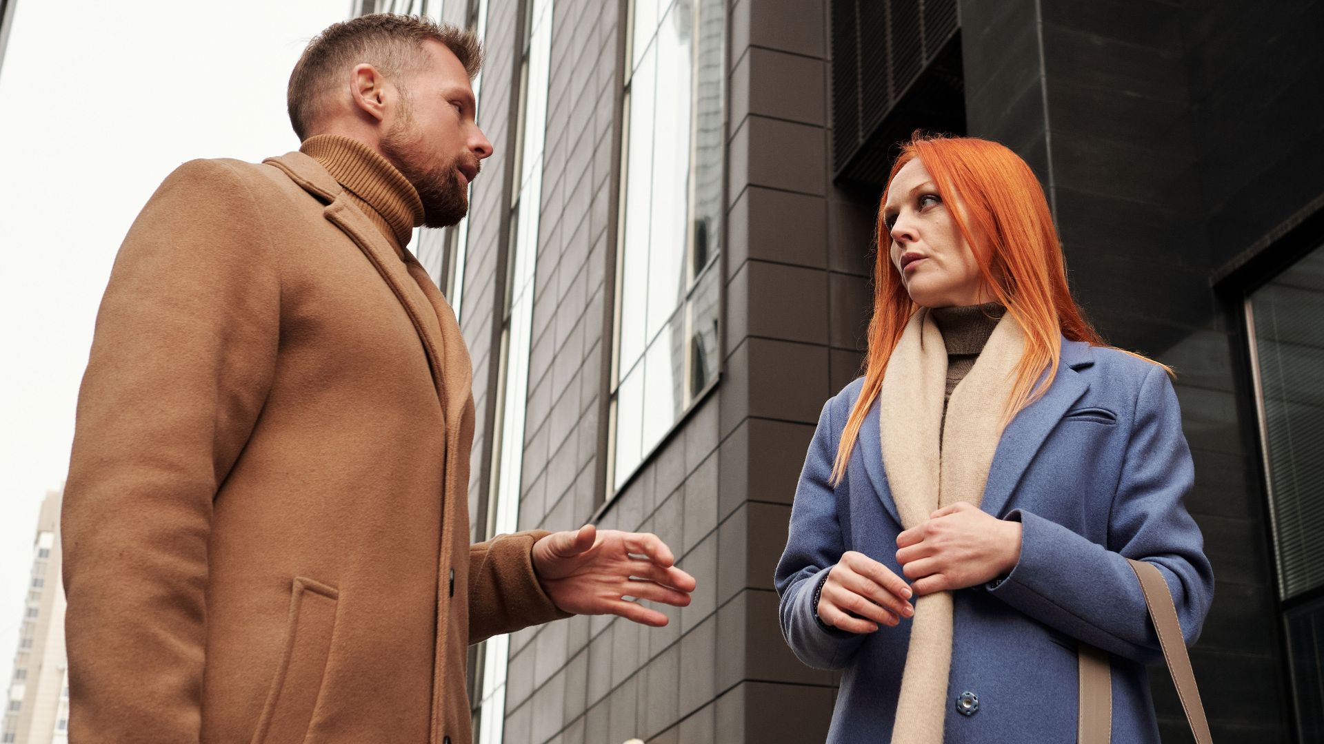 Two adults engage in a conversation outdoors between modern city buildings.