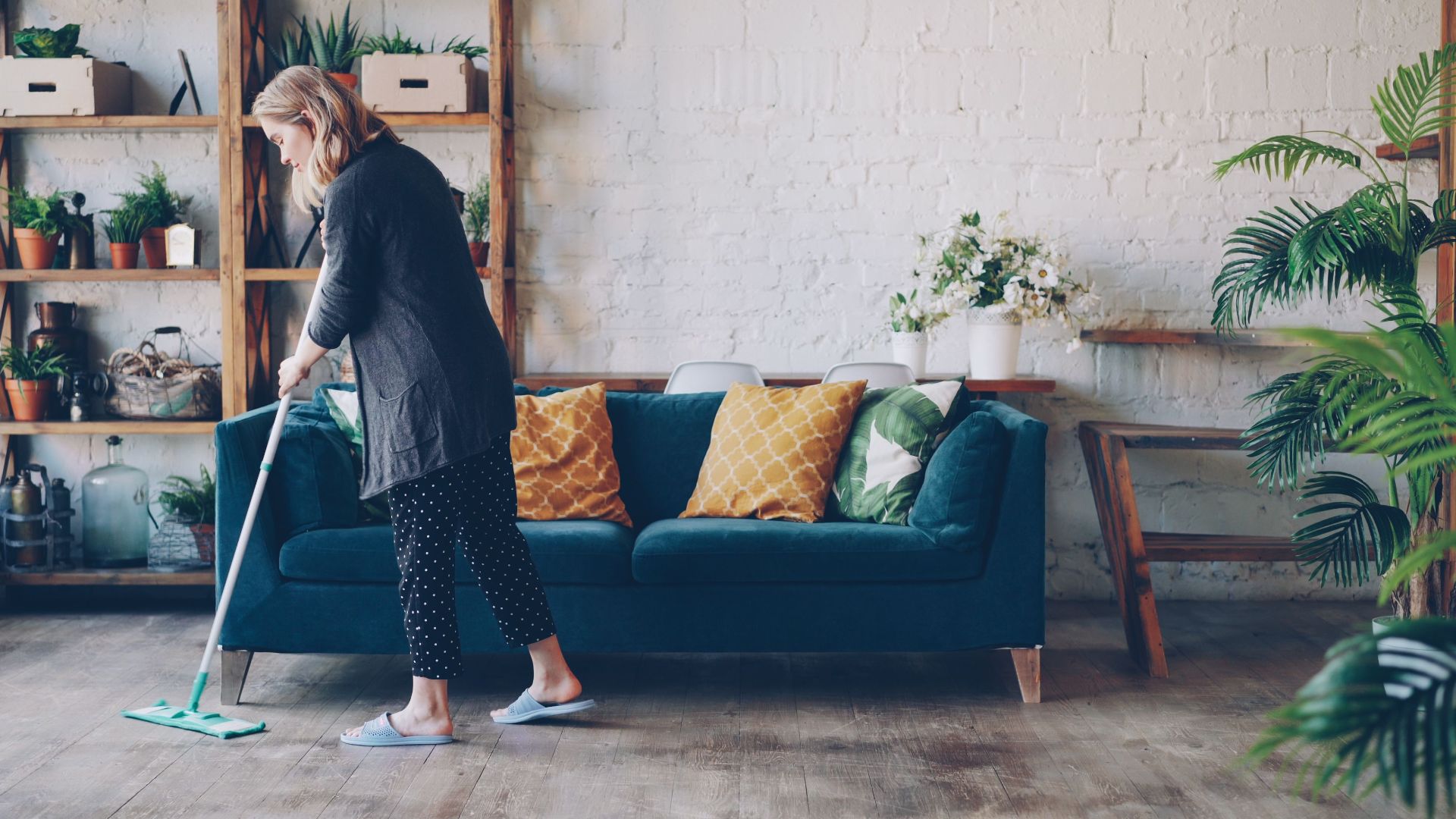 Woman with mop cleaning cozy living room with blue sofa and plants.