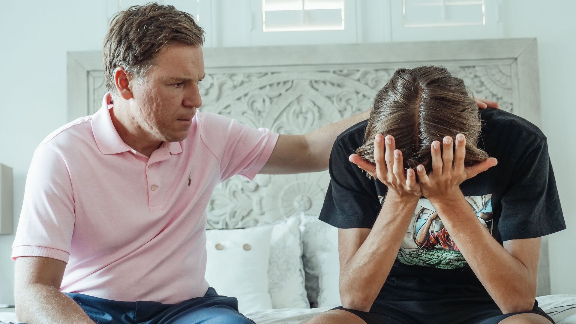 A compassionate father consoles his upset teenage son on a bed indoors.