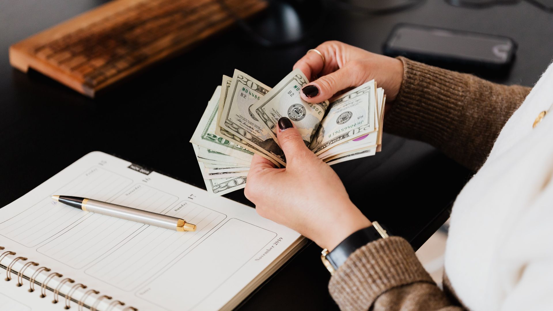 Unrecognizable elegant female in sweater counting dollar bills while sitting at wooden table with planner and pen