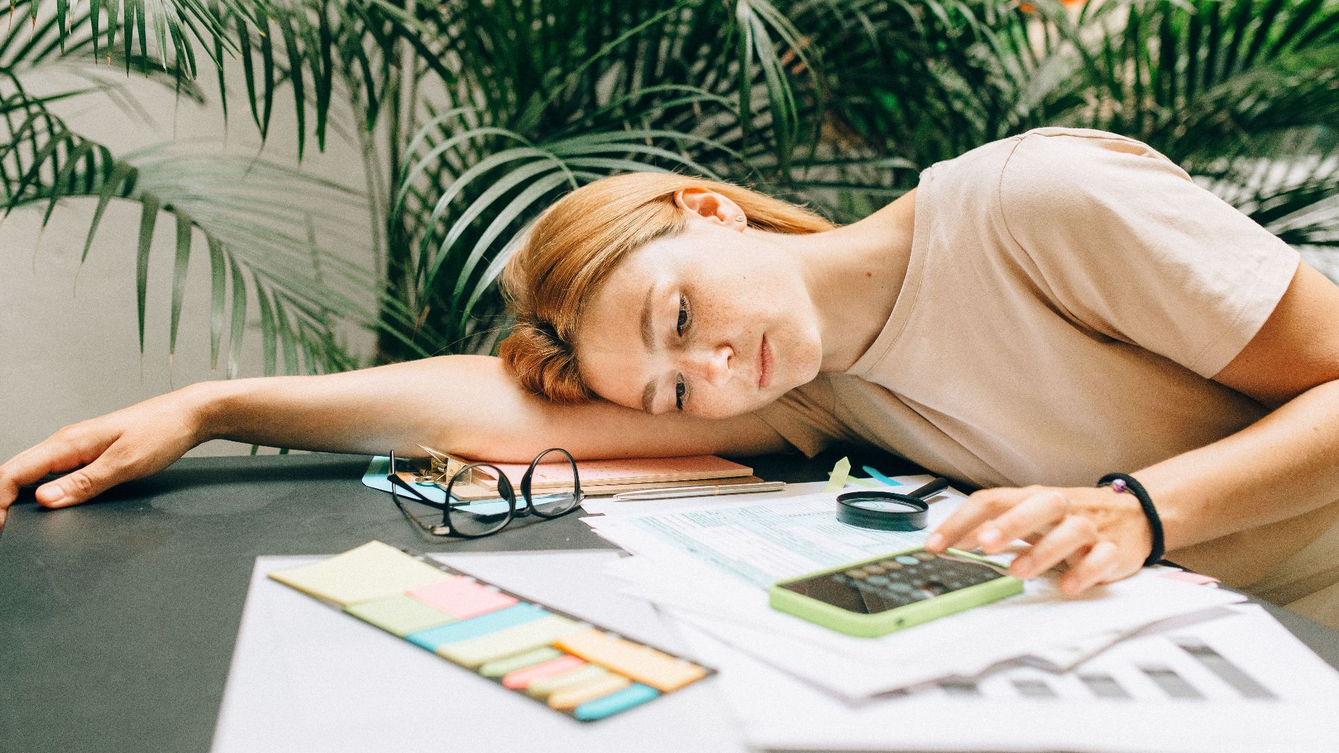 A tired woman calculating her taxes at a desk covered with papers and a smartphone.