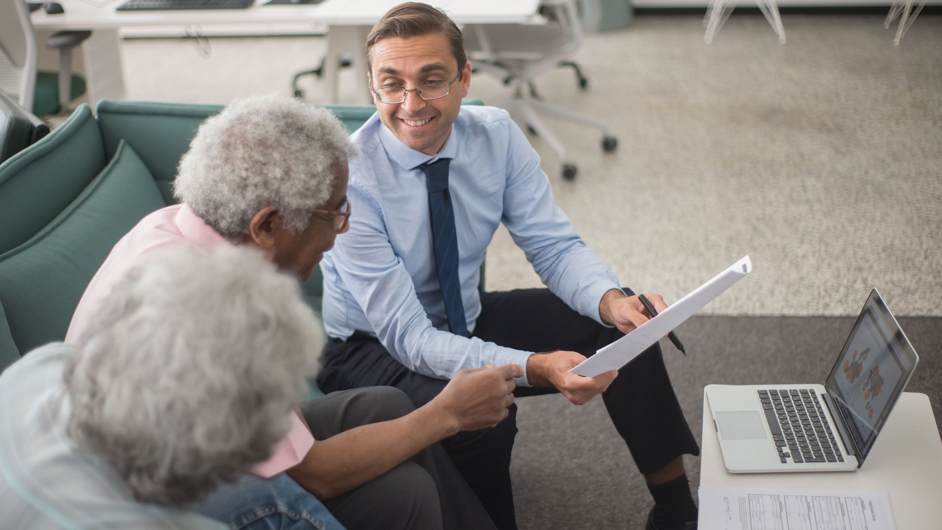 Consultant discussing financial plans with senior clients in a modern office setting, using documents and a laptop.