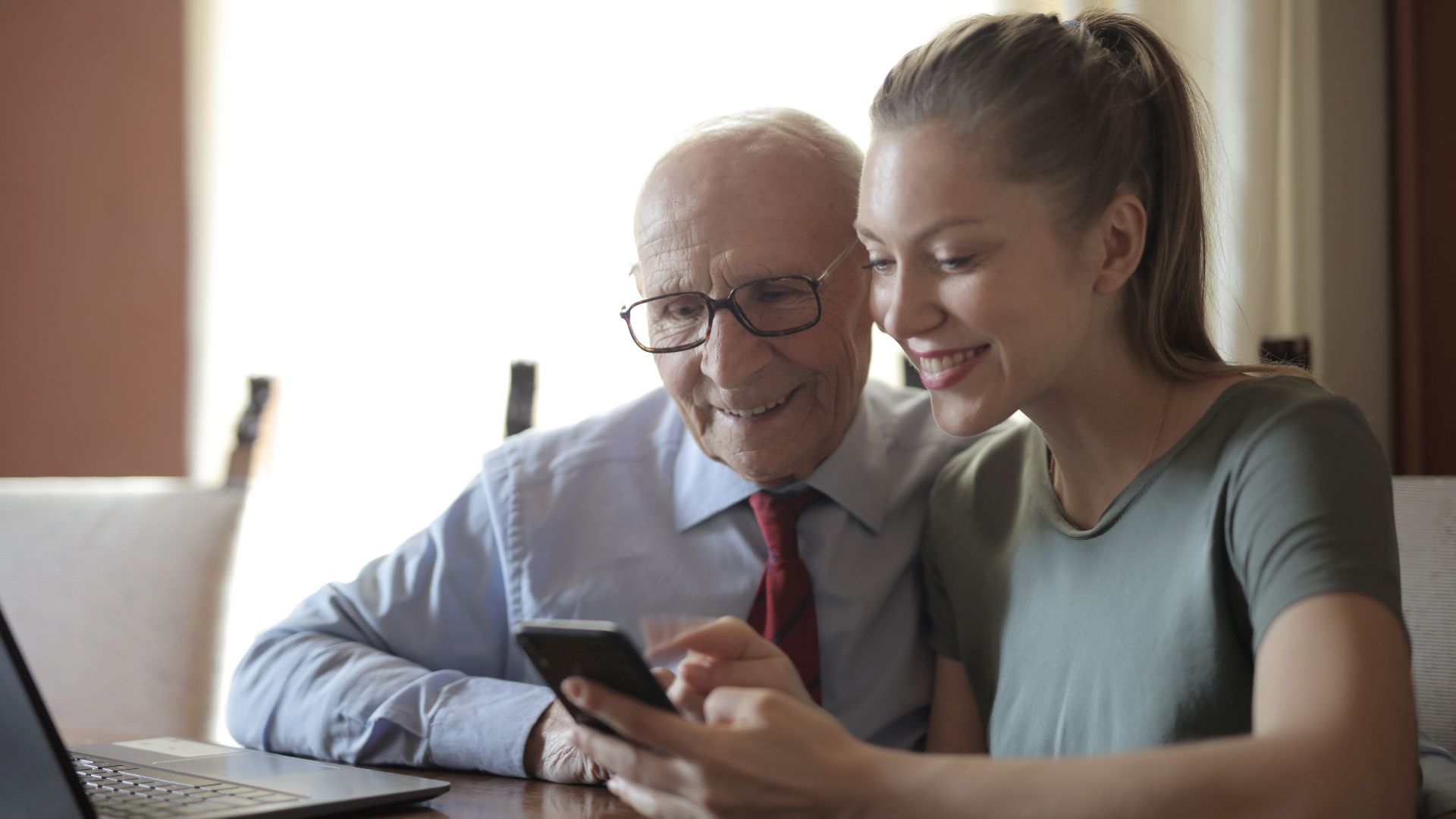 Smiling young woman in casual clothes showing smartphone to interested senior grandfather in formal shirt and eyeglasses while sitting at table near laptop