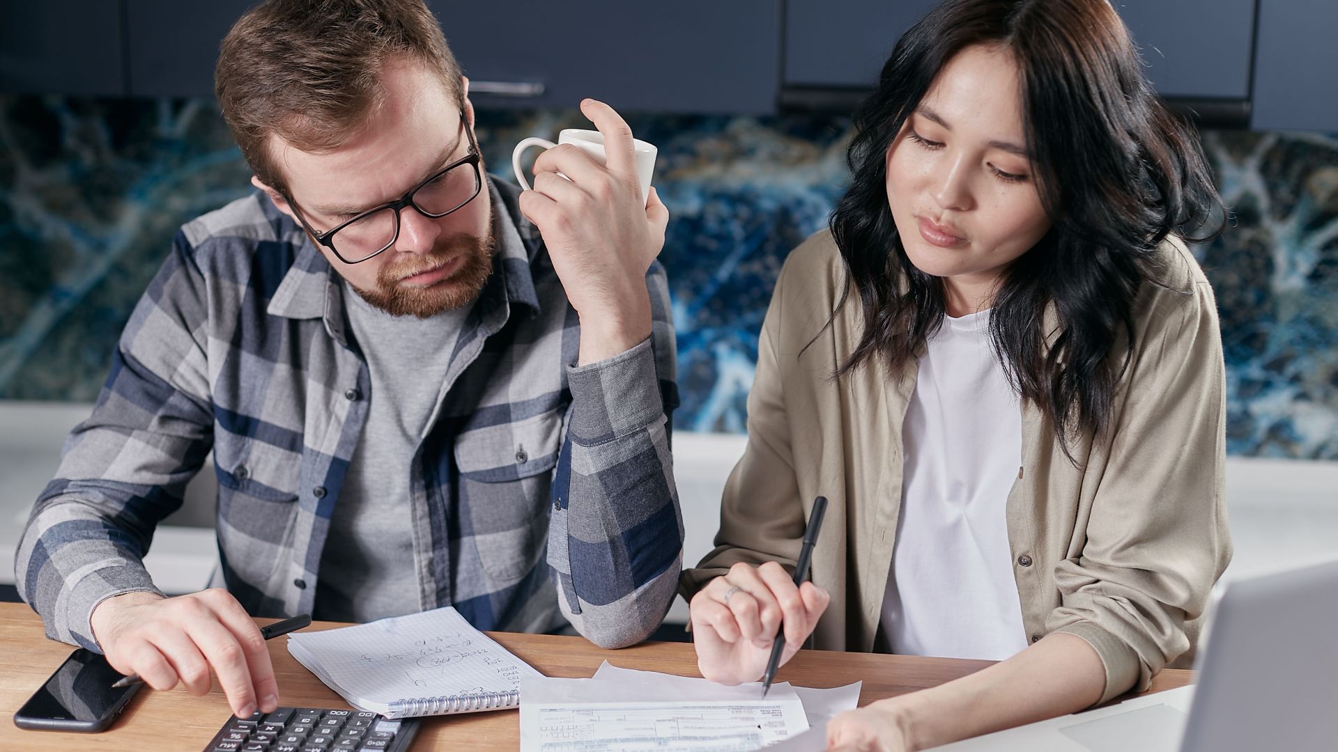 A couple reviewing household bills and budget using a calculator and laptop at their kitchen table.