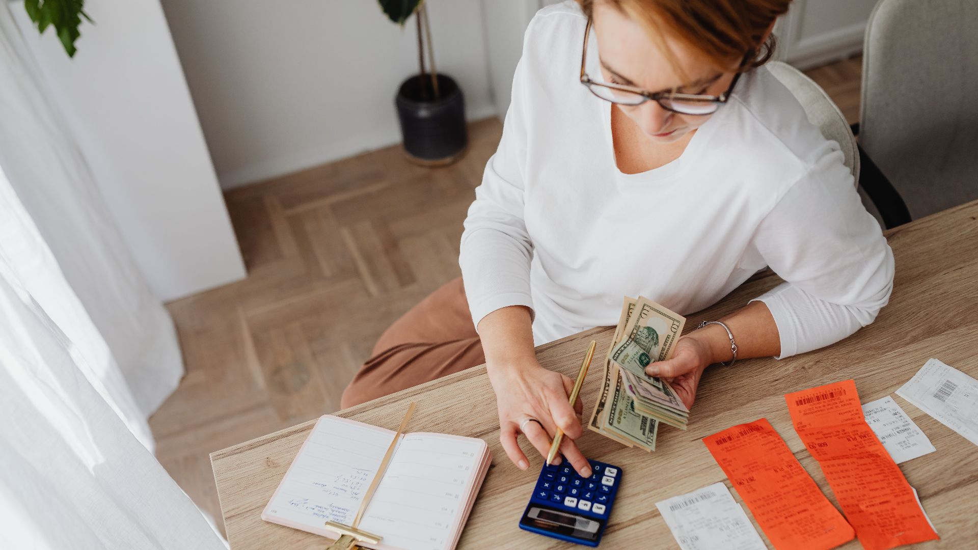 Top view of a woman managing finances with a calculator, cash, and bills in a home setting.