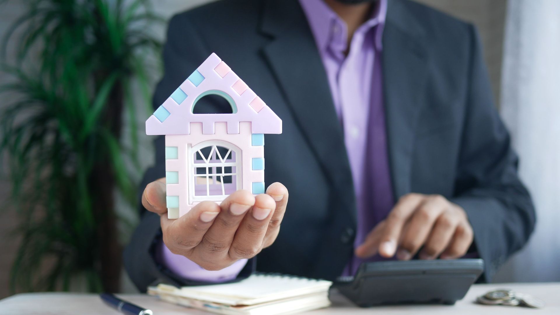 Businessman in suit holds model house, calculates real estate investments.