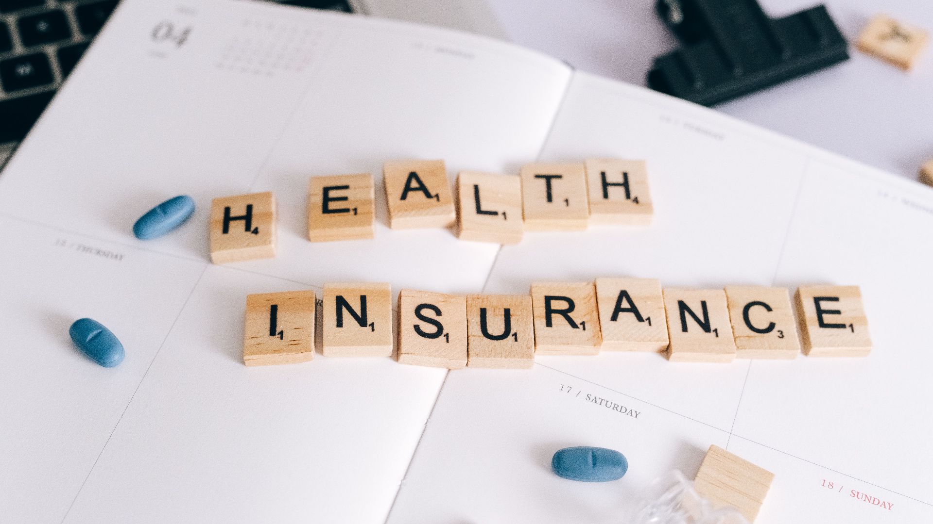 Scrabble tiles spelling 'Health Insurance' on planner with pills and laptop, symbolizing healthcare planning.