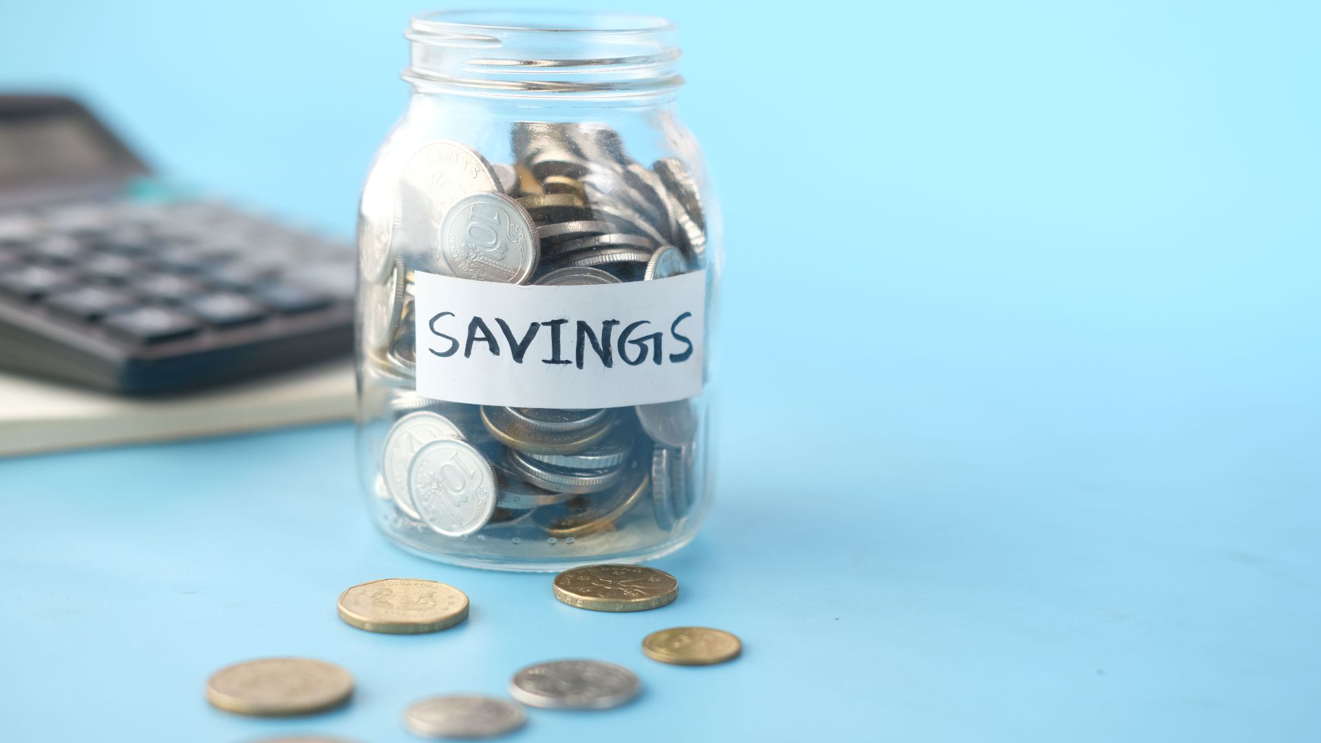 Glass jar labeled 'Savings' filled with coins, beside a calculator on a blue background.