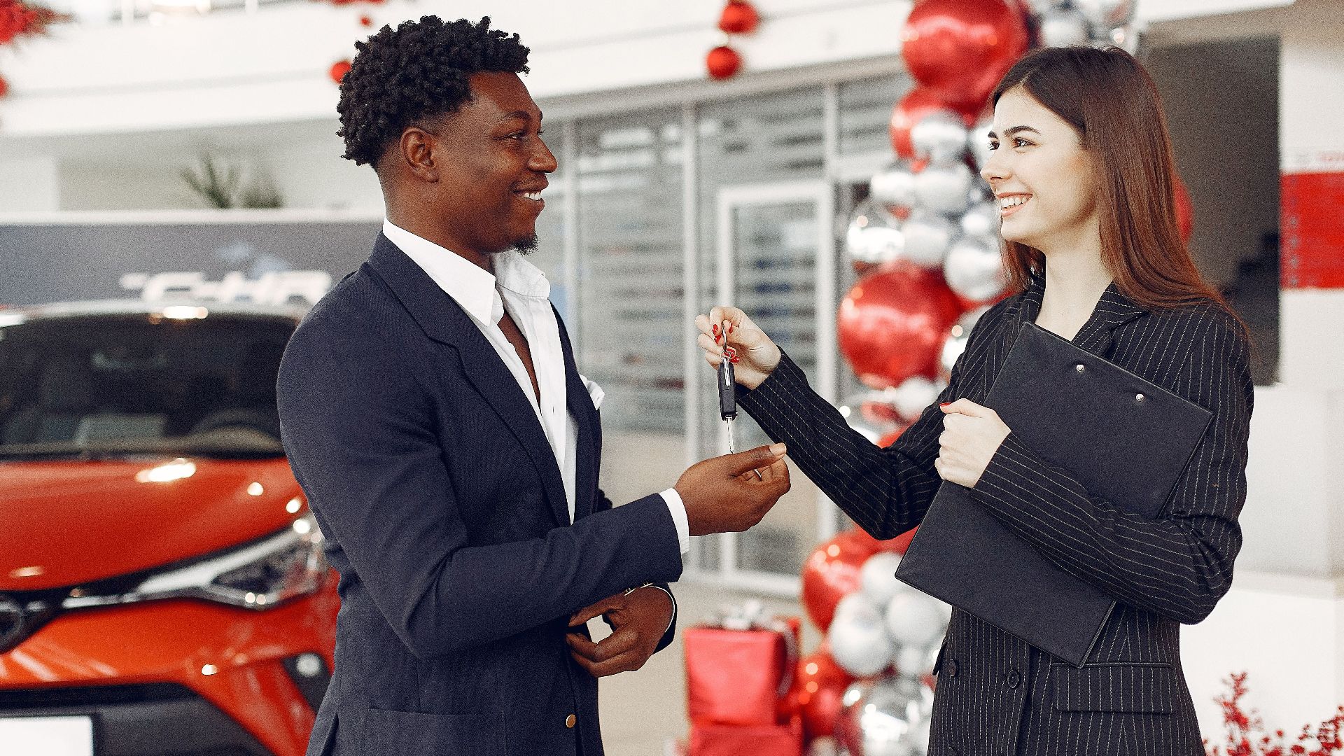 Side view of happy positive female dealer in formal classy wear passing car keys to excited African American customer in trendy suit in car showroom
