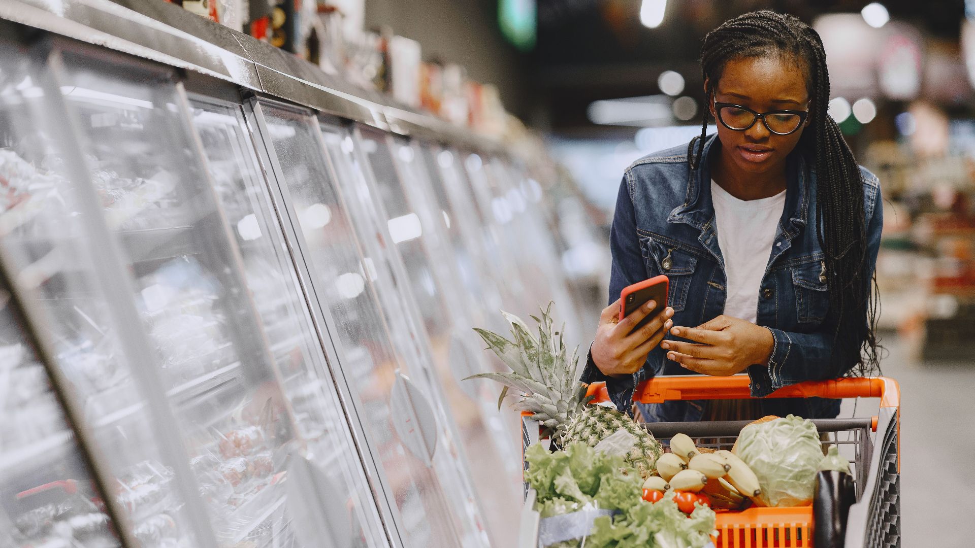 Woman in a grocery store using smartphone while pushing a shopping cart filled with groceries.