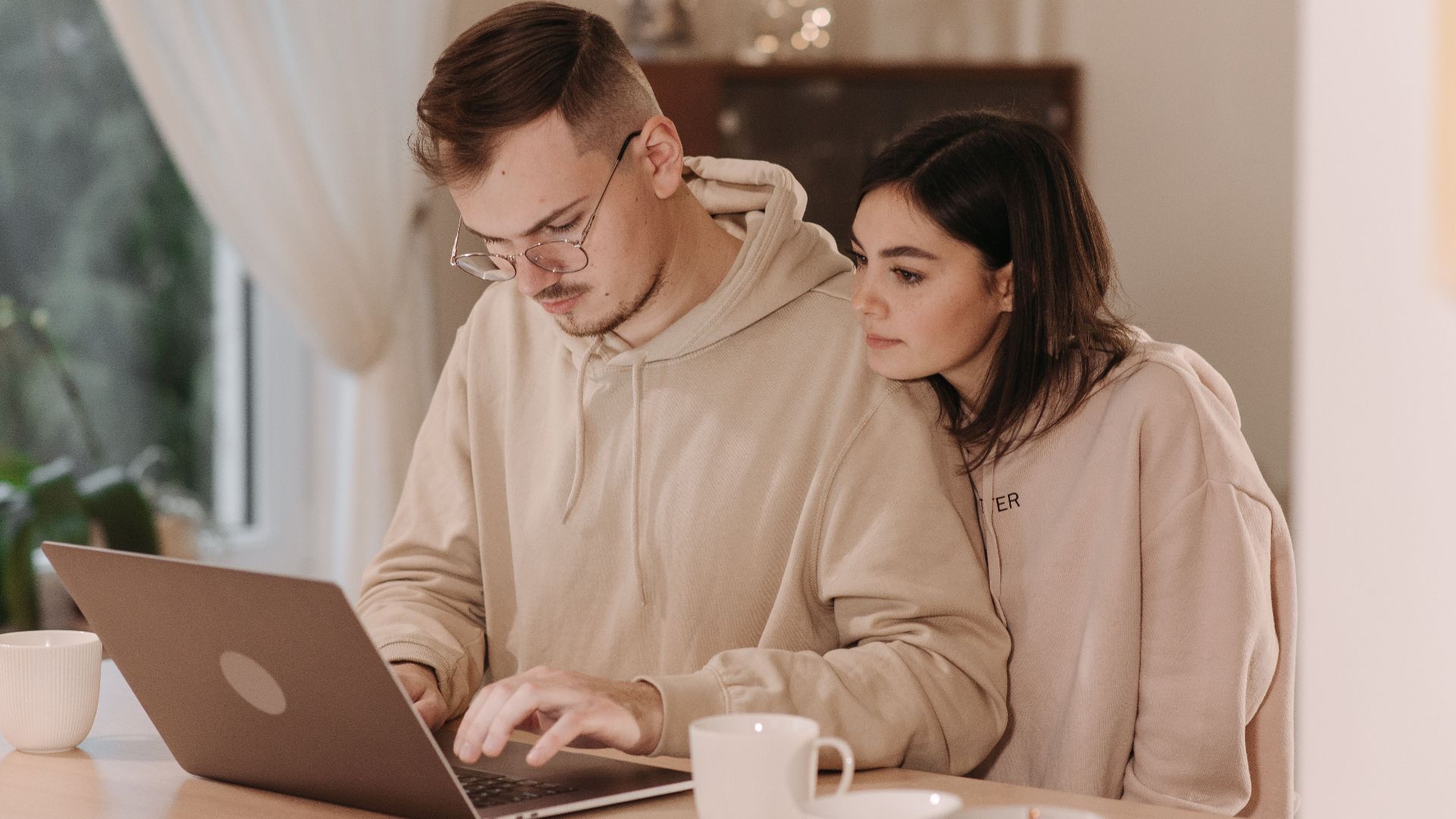 Young couple in hoodies using a laptop at home with coffee cups, embracing remote work lifestyle.