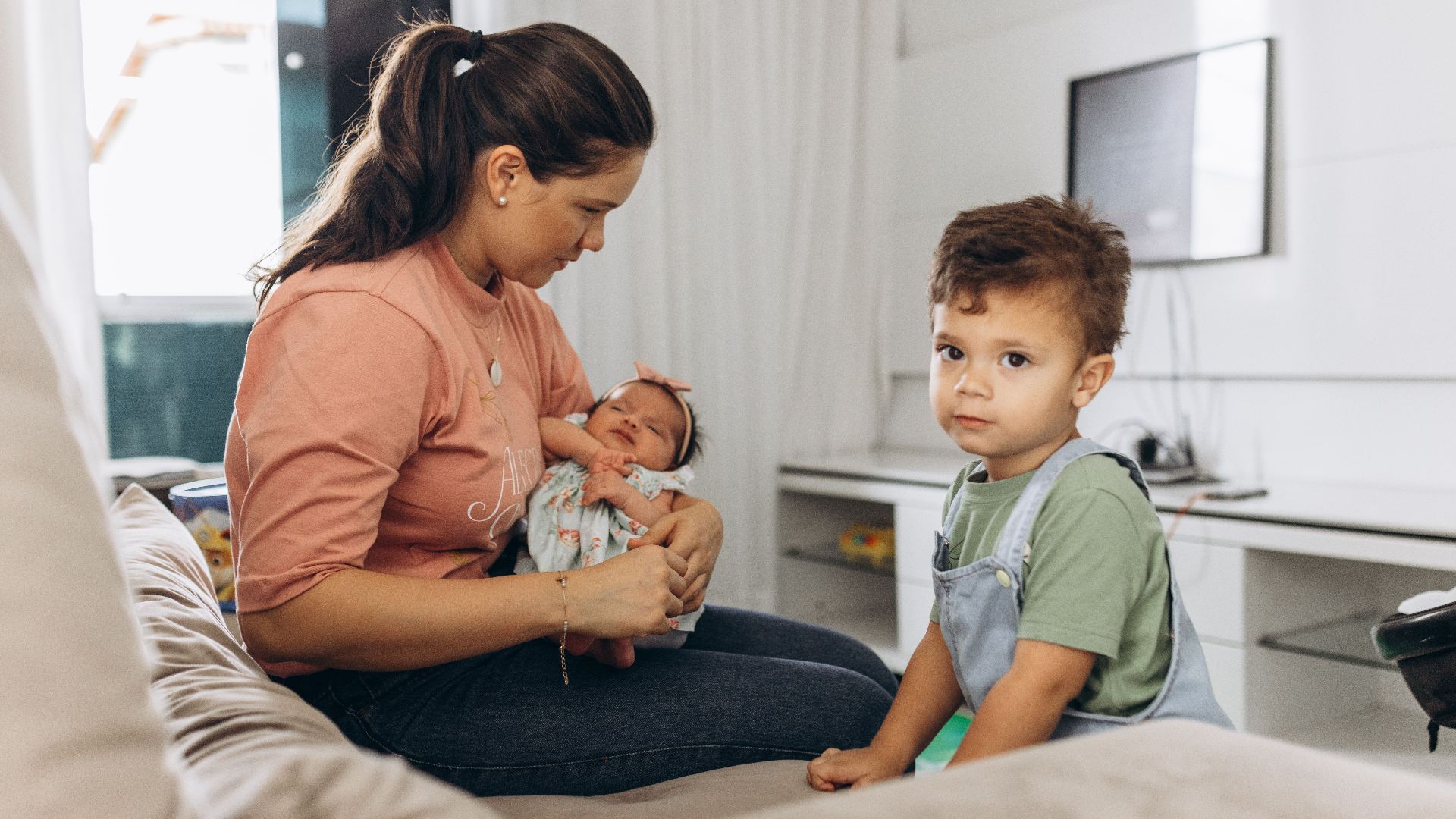 A mother sitting on a sofa with her baby and young son in a bright living room setting.