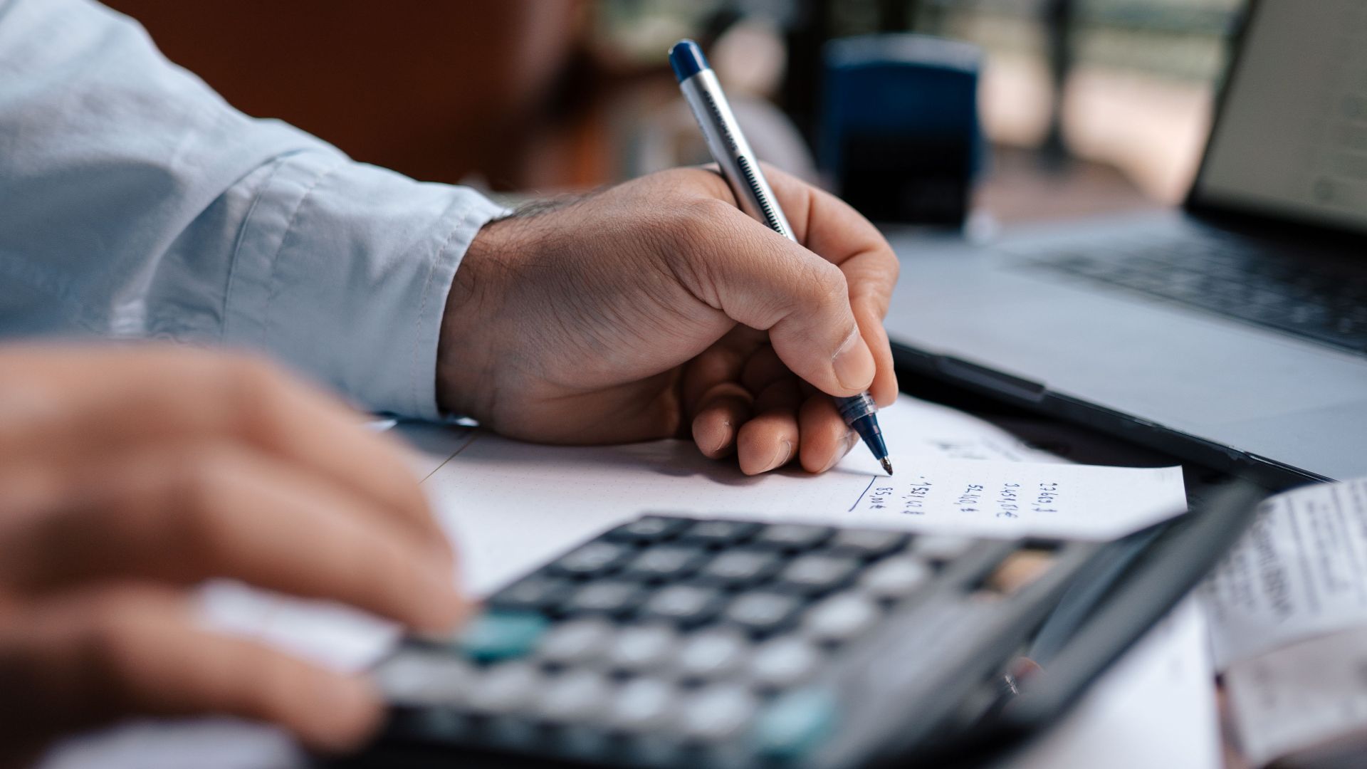 A person calculating finances with a calculator and pen on a desk indoors.