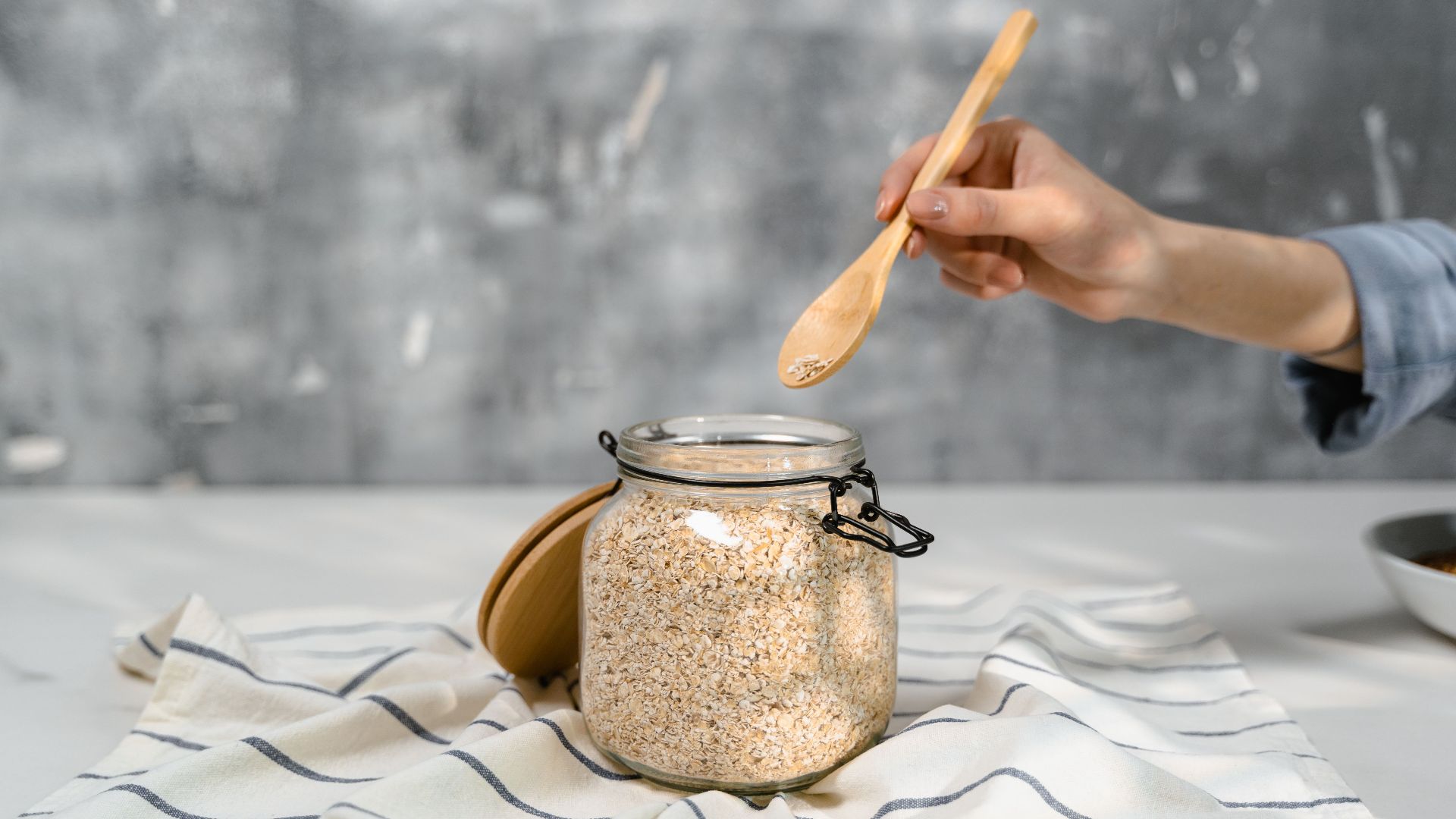 Hand holding a wooden spoon over a glass jar filled with oats on a striped tablecloth.