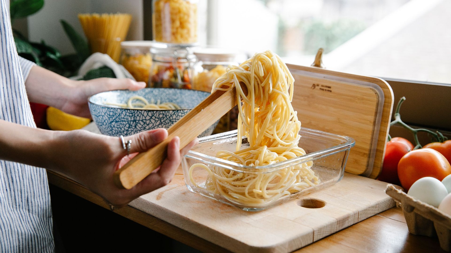 Crop anonymous female cook putting spaghetti from bowl into glass container using wooden tongs while cooking at counter with assorted ingredients