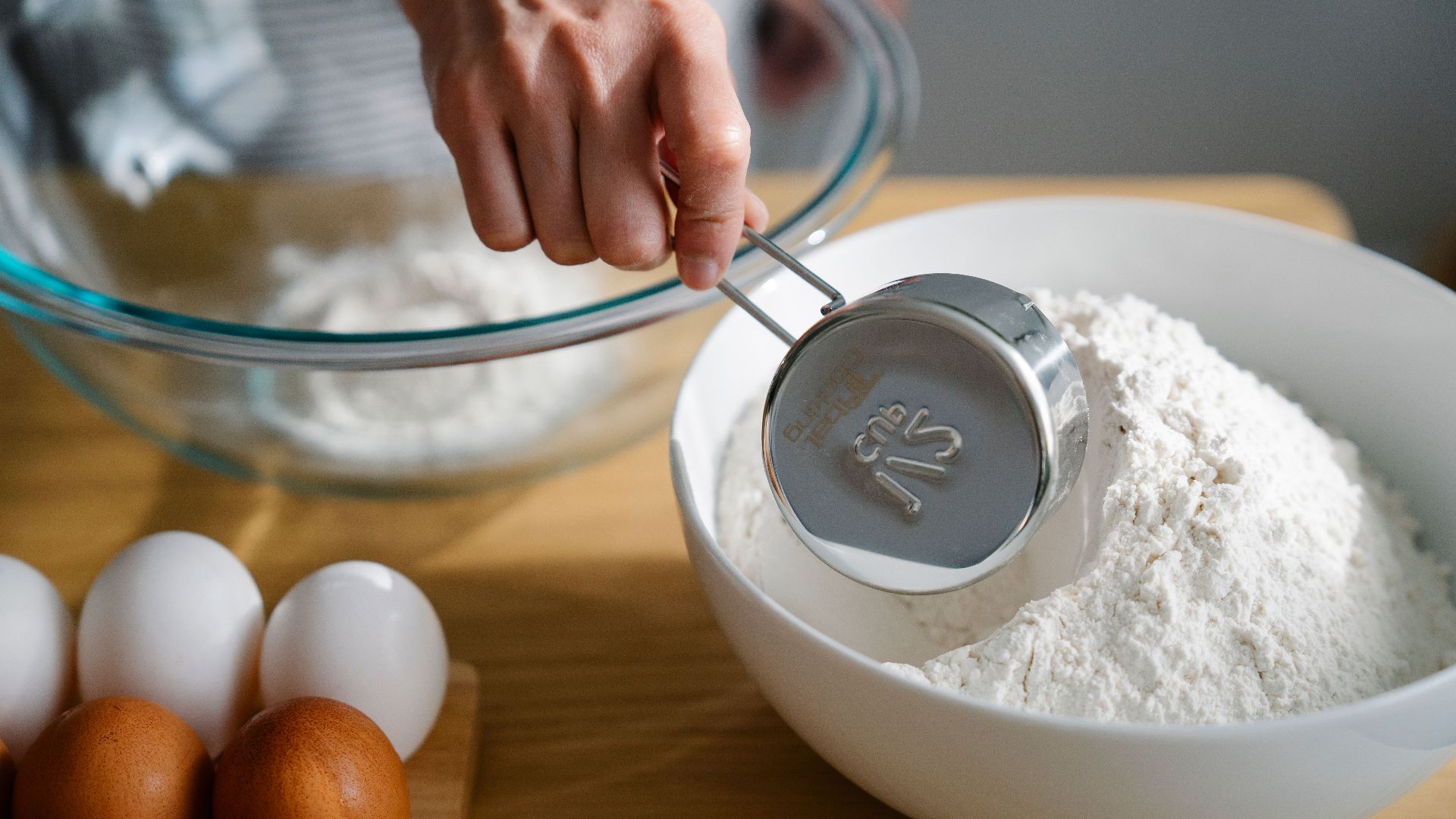 Close-up of a hand measuring flour in a bowl, ready for baking with eggs nearby on the kitchen counter.