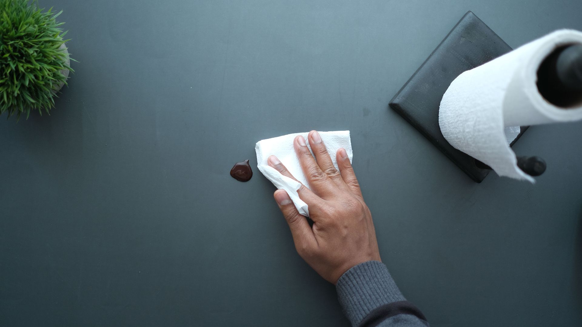 A hand wipes a dark surface with a tissue near a paper roll and plant.