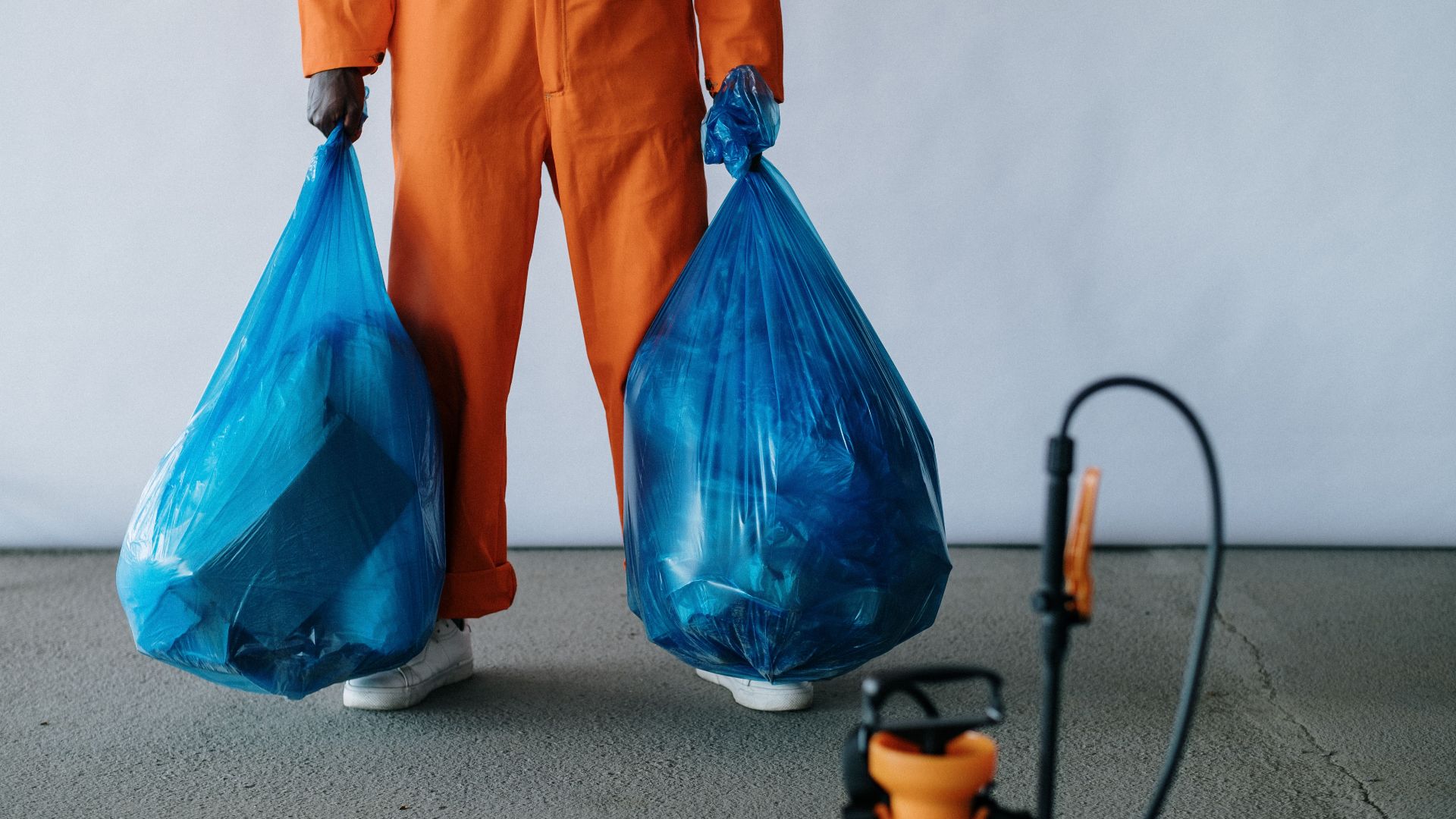 Sanitation worker in orange overalls holding blue trash bags indoors for waste management.