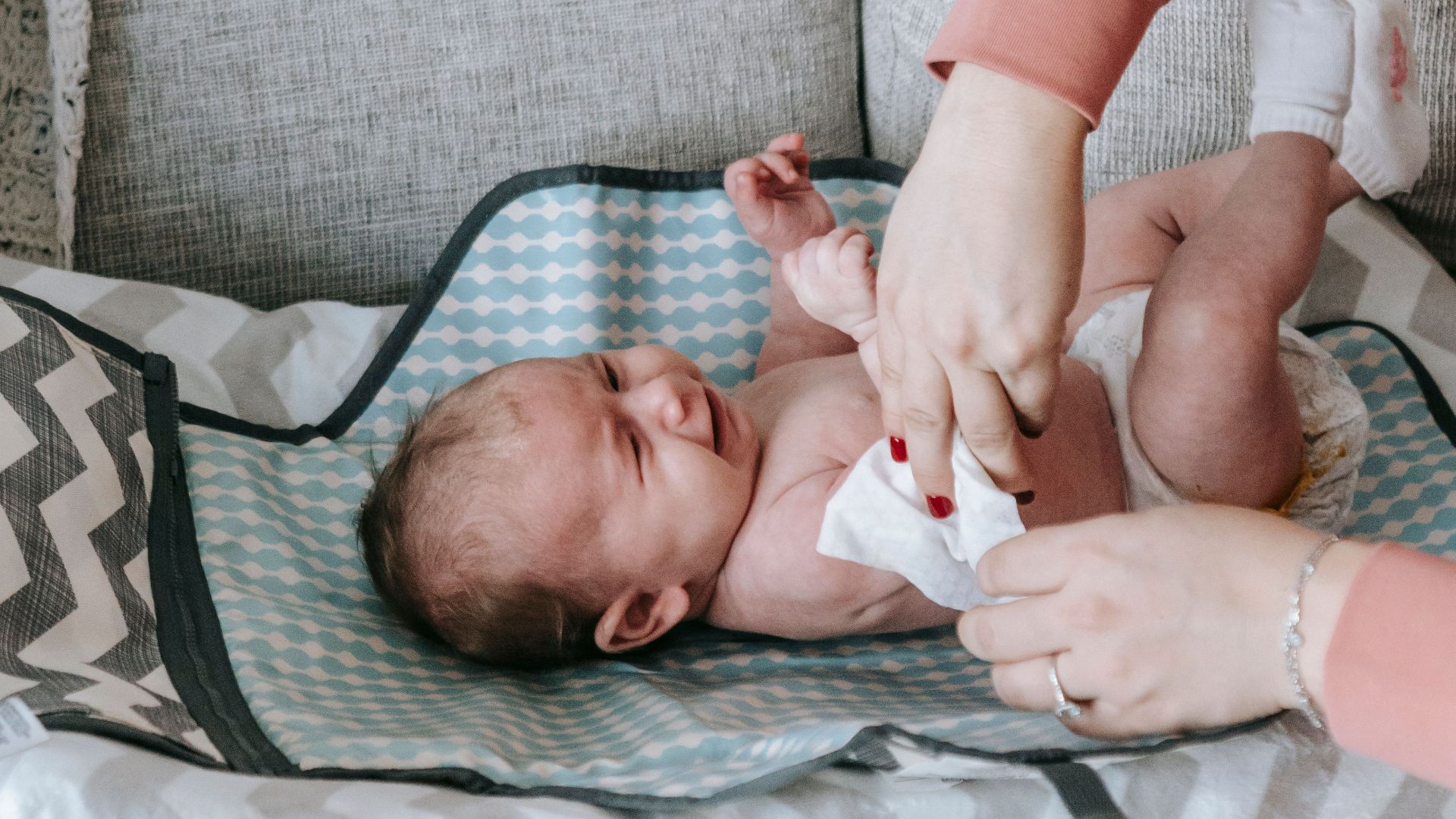 A caregiver gently changes a crying baby's diaper on a sofa at home.