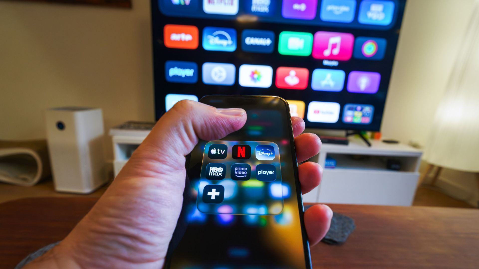 Close-up of a hand holding a phone displaying streaming apps in front of a TV with multiple app icons.