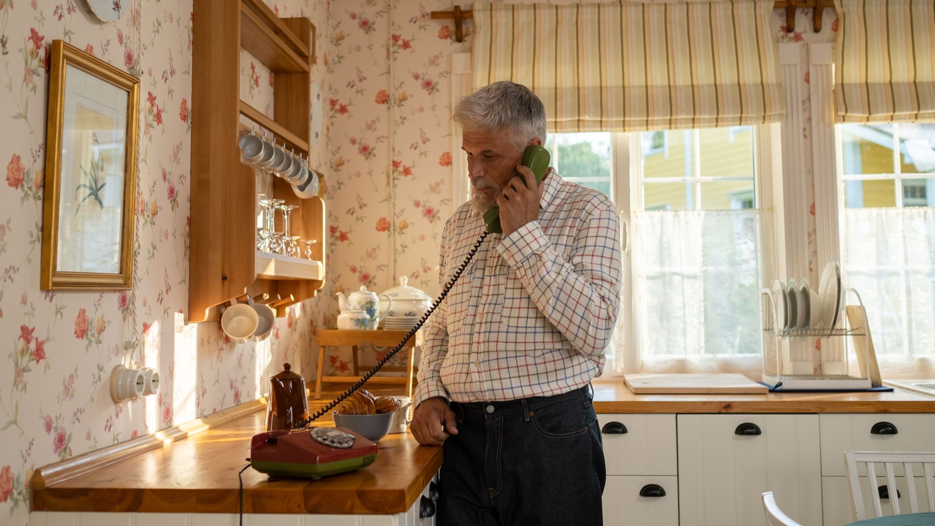 Senior man talks on a vintage rotary phone in a retro-styled kitchen with floral wallpaper and classic decor.