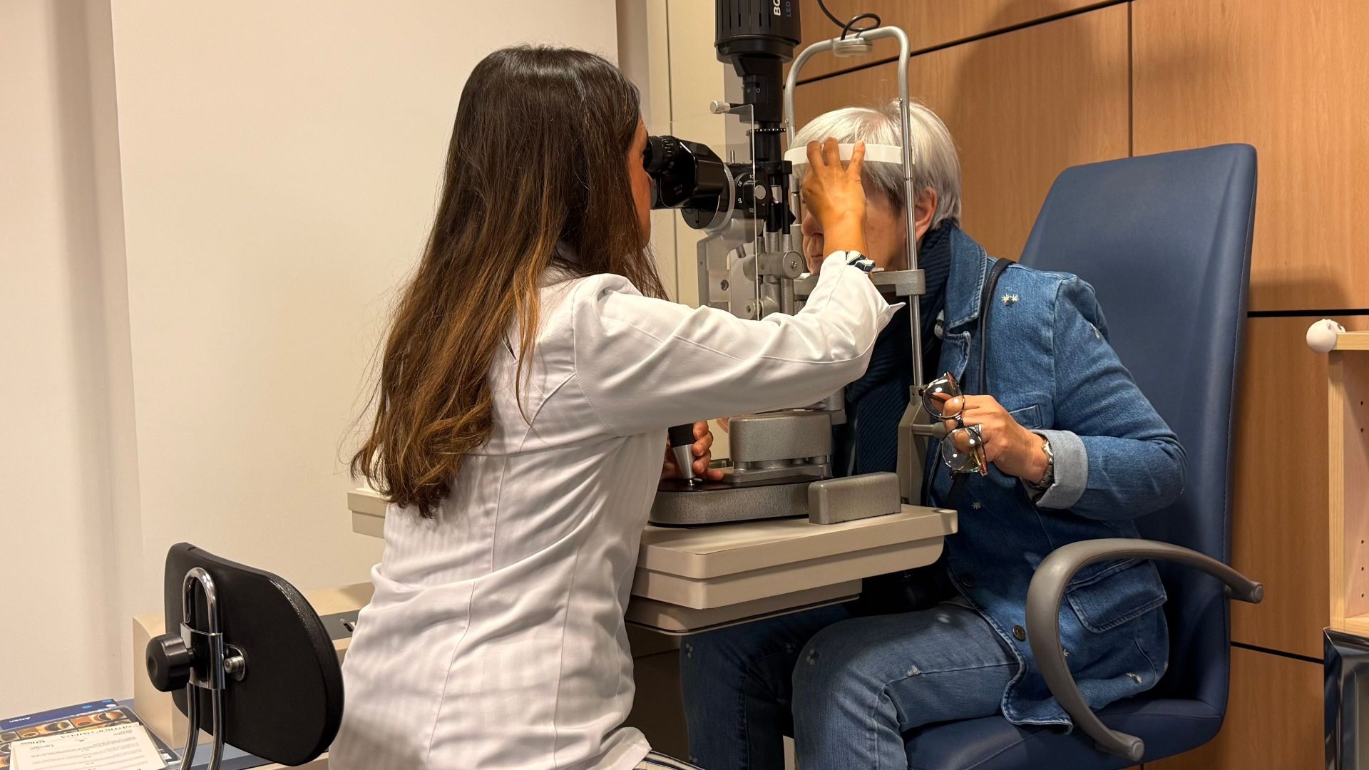 Optometrist performing an eye exam with a slit lamp in a well-lit examination room.